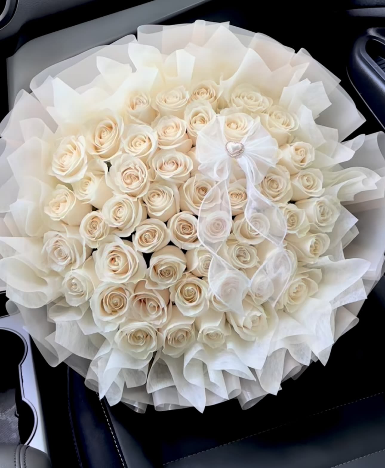 Pure Love - A breathtaking round bouquet of creamy ivory roses, artfully arranged and wrapped in layers of sheer white tulle. Finished with a delicate chiffon bow adorned with a heart detail, this arrangement embodies elegance, purity, and refined romance.