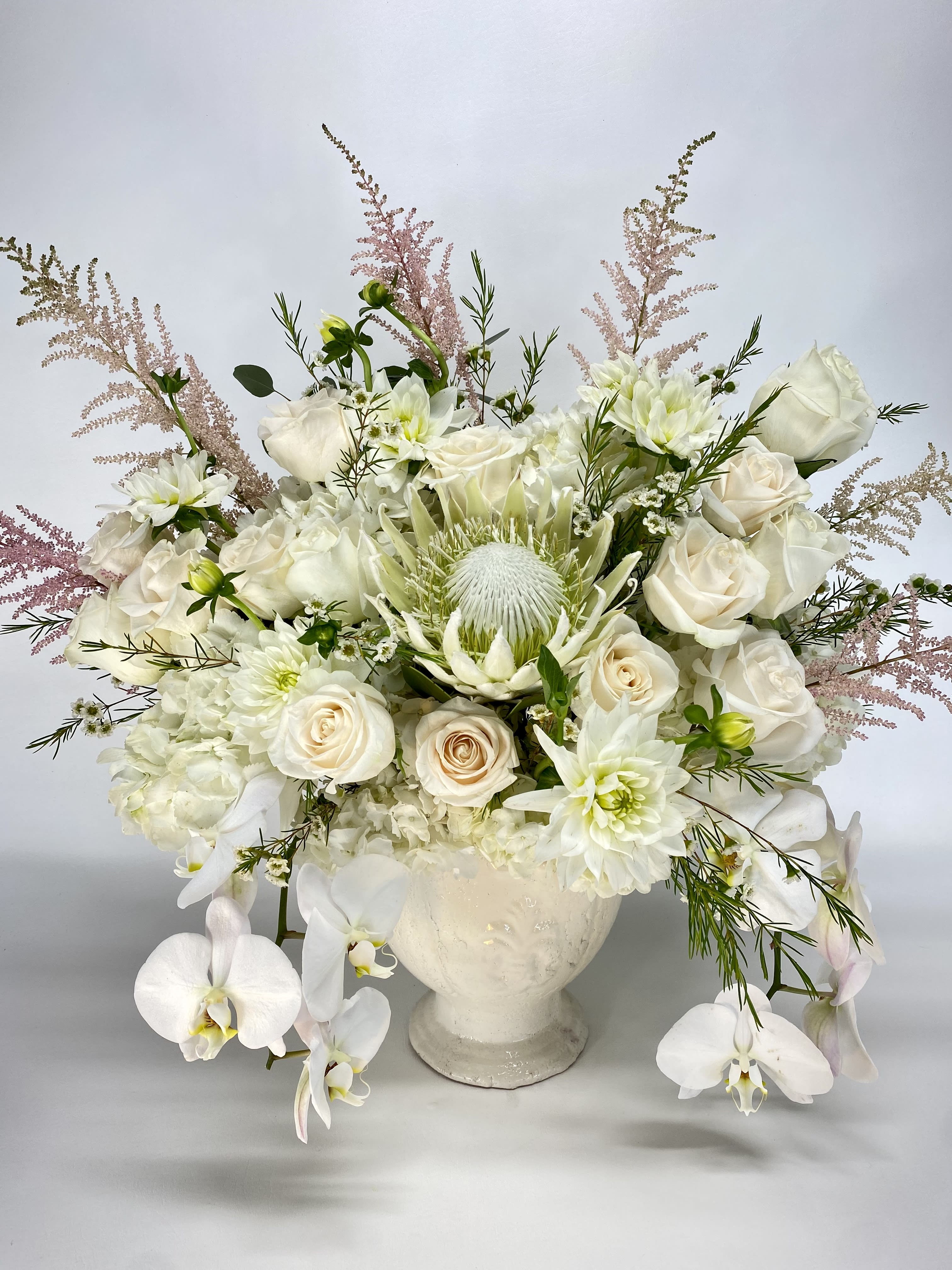 White Royalty  - Very elegant all white floral arrangement in a rustic impressive footed vase. Flowers include Hydrangeas, Roses, Dahlias, king Protea and Orchids.