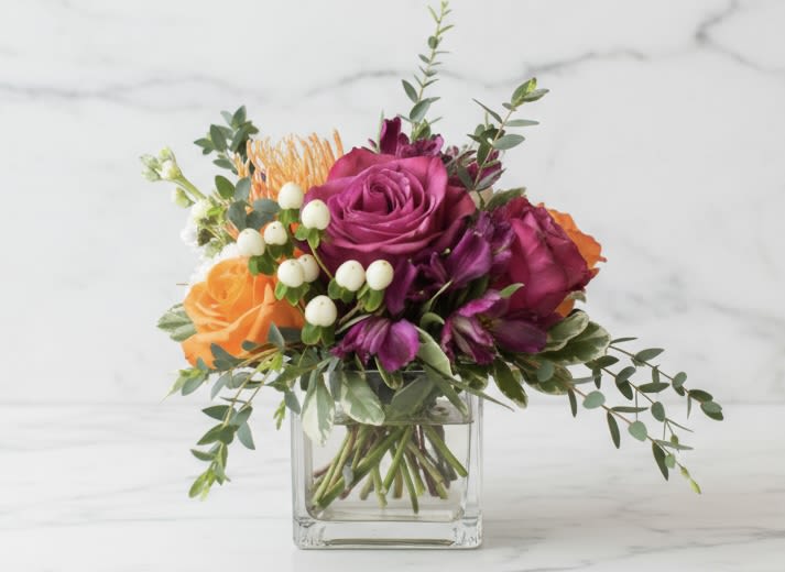Nirvana - low arrangement - Low Arrangement in a Clear Glass Cube of jewel tone roses, pin cushion, alstromeria, stock, mums, berries and flowing greens.