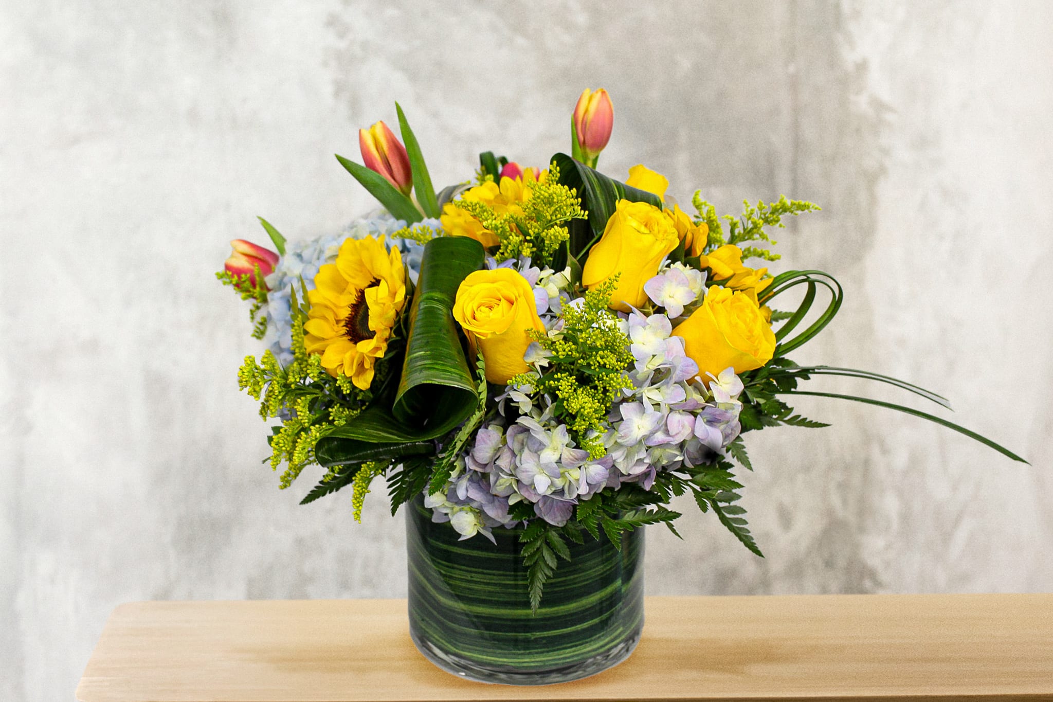 Roses, Tulips, &amp; Sunflowers - This Modern compact arrangement is designed with Hydrangea, Yellow Roses, Sunflowers, Tulips, Solidago, and tropical leaves. 