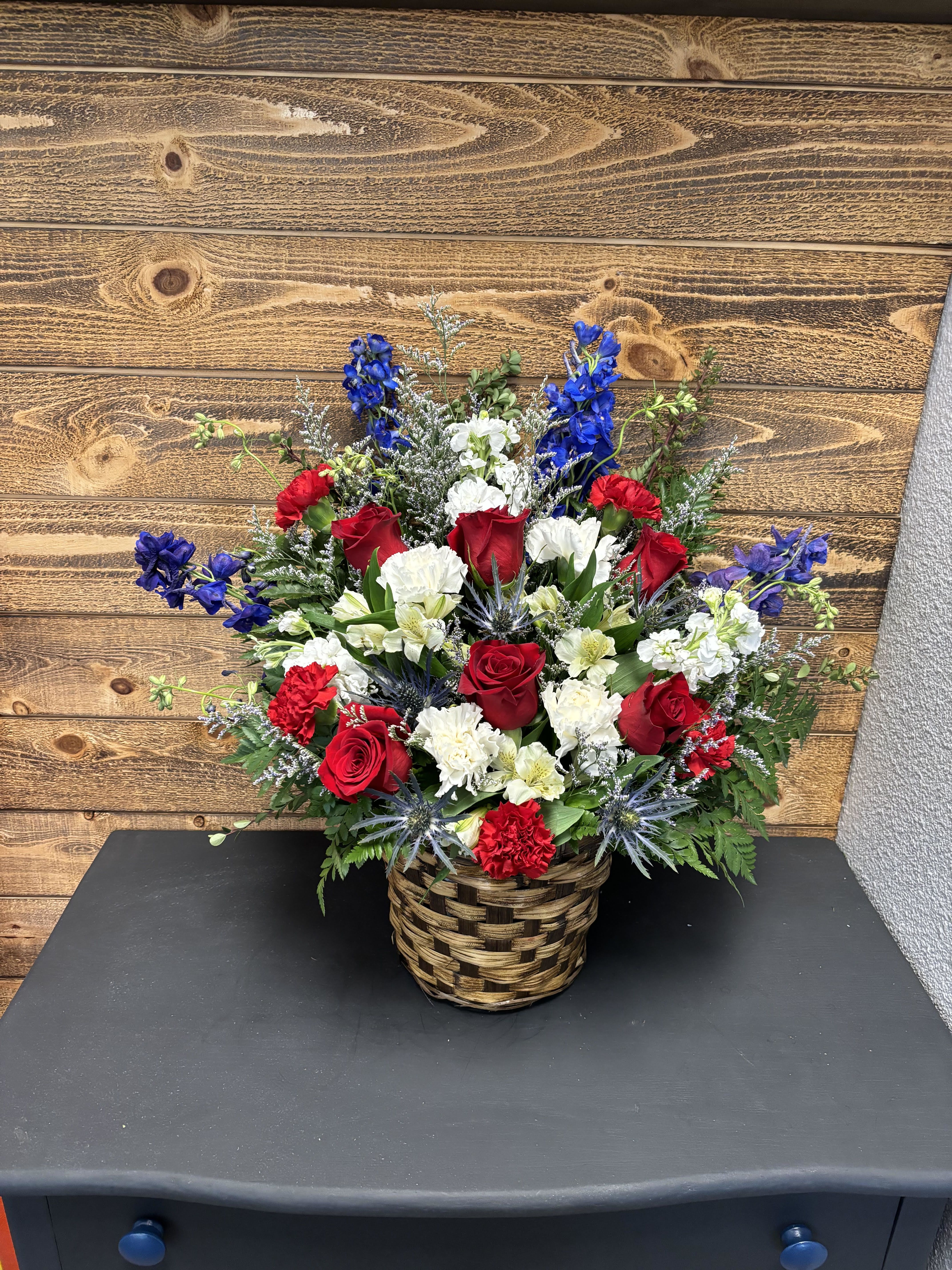 Forever Remembered by Anderson Florist - A red, white, and blue tribute funeral basket featuring red roses, blue delphinium, carnation and white stock. Fitting for any type of funeral ceremony.