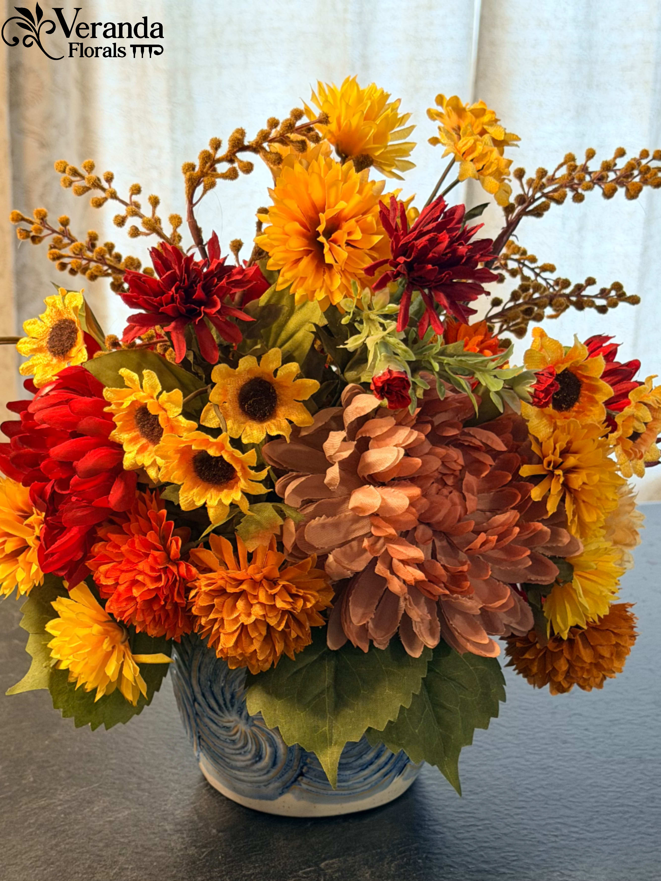 The Cerulean Sun | Permanent Botanical Arrangement - The Design Brighten any corner of your home with The Cerulean Sun, a vibrant arrangement that celebrates the harmony of warm and cool tones. This design features a lush mix of golden-yellow sunflowers, fiery red mums, and rich terracotta zinnias. We’ve added height and airy texture with delicate seeded sprigs that reach upward, mimicking the natural growth of a wildflower garden.  The dense, rounded composition provides a full, 360-degree view, making it an ideal choice for a centerpiece where beauty is needed from every angle.  The Vessel The arrangement is housed in a stunning blue and white chinoiserie-inspired vessel. The ceramic pot features a swirling, textured wave pattern in deep cobalt, providing a crisp, classic contrast to the vivid oranges and reds of the florals. This combination of traditional pottery and bold botanicals makes it a perfect fit for a grandmillennial or classic traditional interior.