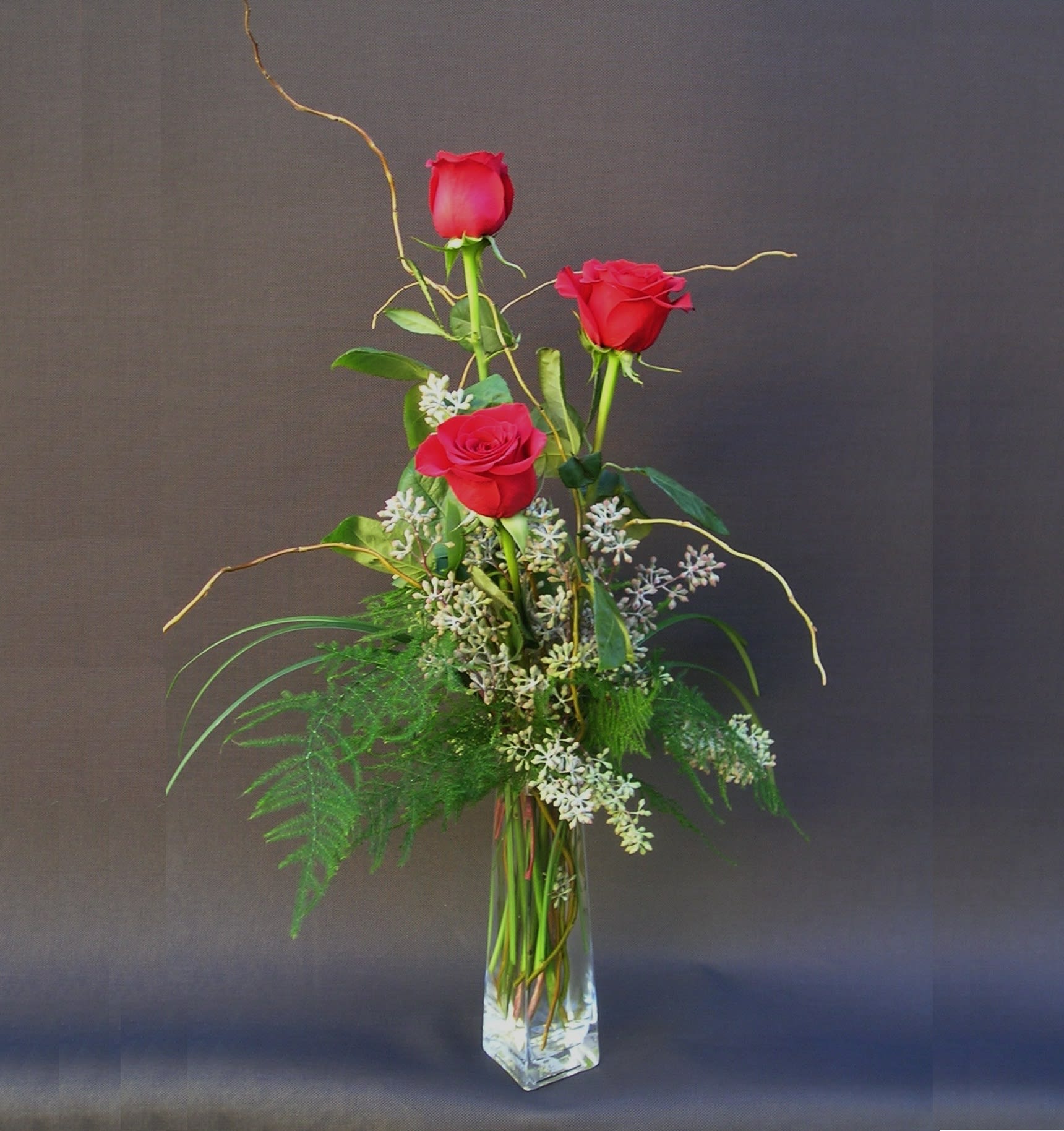 Red Rose Bud vase  - Featuring three vibrant red roses, elegantly arranged with complementary greenery and subtle filler accents in a clear glass vase. This classic and minimalist design emphasizes the beauty and symbolism of the roses themselves. 
