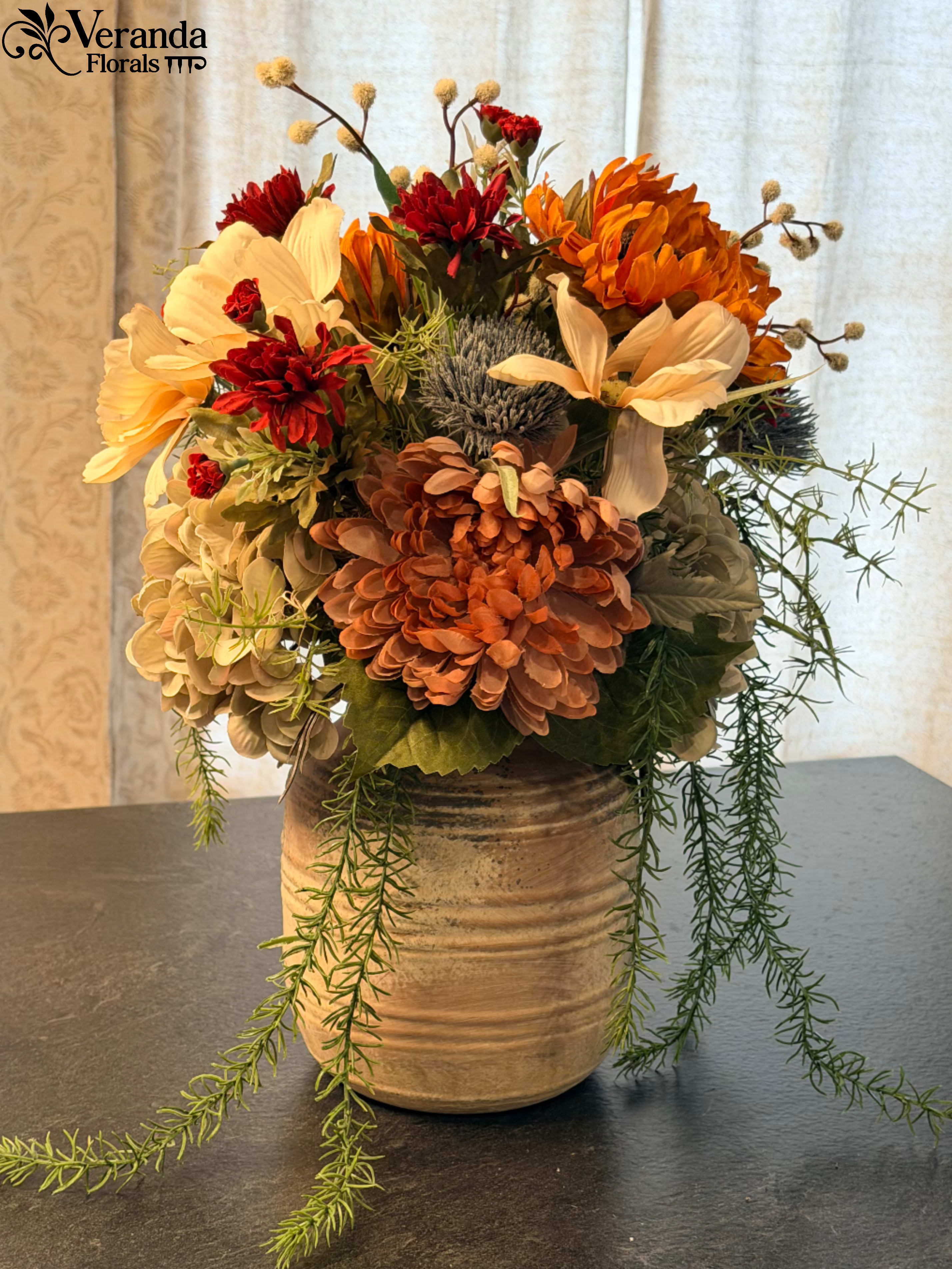 The Terra &amp; Vine | Artisan Permanent Botanical - The Design Bring a touch of the outdoors in with The Terra &amp; Vine. Designed to be a staple of your home decor, this arrangement focuses on organic textures and a sophisticated, earth-toned palette. We’ve paired muted terracotta dahlias and deep burgundy mums with creamy, sun-washed blooms to create a look that feels both grounded and elevated.  The "wild" element of this design is the cascading rosemary-style greenery, which spills naturally over the side of the vessel, giving the arrangement a fresh, "just-cut" movement that breathes life into any room.  The Vessel The arrangement is anchored in a heavy-ribbed stoneware vase. Its weathered tan glaze and horizontal ridges offer a tactile, artisan feel that sits perfectly in a modern farmhouse, a grandmillennial library, or a minimalist entryway.