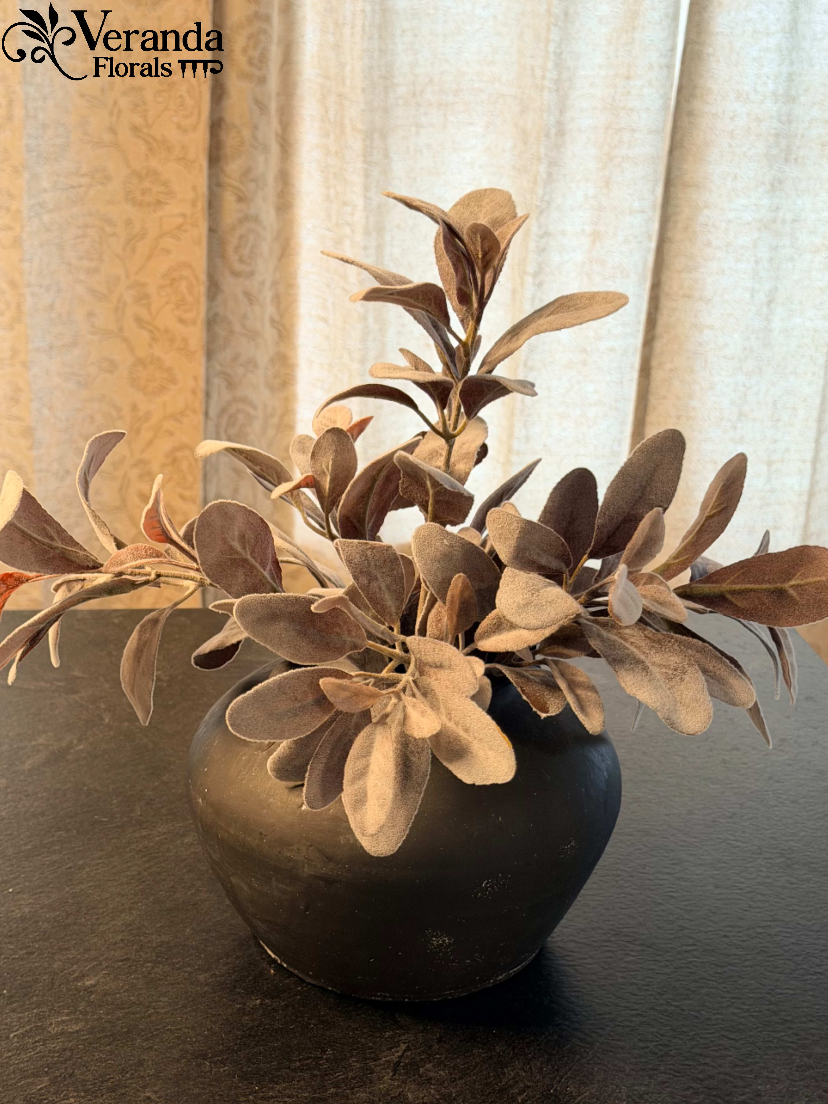 The Velvet Lamb’s Ear Urn - Understated. Tactile. Timeless Bring the soft, silvery elegance of a classic English garden indoors. This permanent botanical features remarkably lifelike Lamb’s Ear, known for its signature "felted" texture and muted sage-and-silver palette.  Unlike traditional bright greenery, the dusty tones of the Lamb’s Ear provide a sophisticated, neutral accent that complements any season. Each stem is carefully placed in a low-profile, matte charcoal artisan vessel, creating a moody, modern contrast against the soft, velvety foliage.