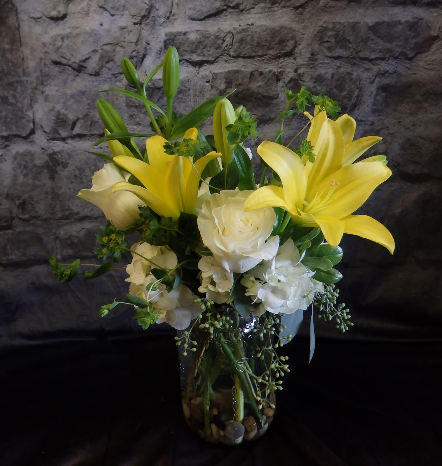 Buttercup - Bright and cheerful, Buttercup radiates warmth with sunny yellow lilies, creamy white roses, and soft, fluffy Hydrangea arranged in a clear jar vase and natural river stones. This uplifting bouquet brings joy and light to any space, perfect for a just-because gesture, celebration, or gentle remembrance. 