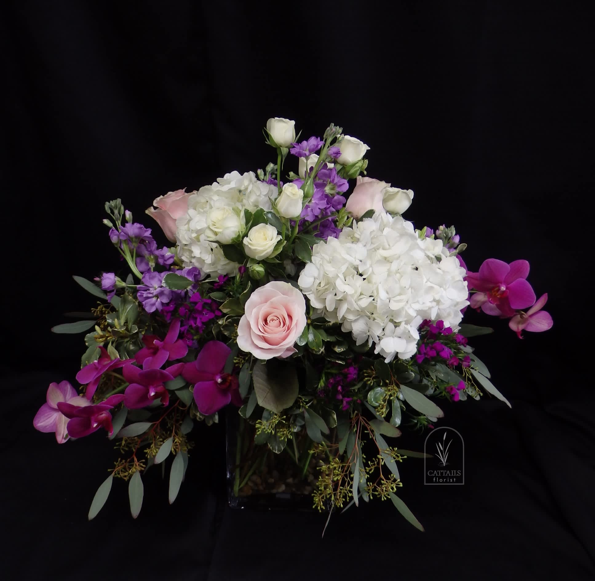 Cosmopolitan - The "Cosmopolitan" flower arrangement features a sophisticated mix of soft blush roses, creamy white hydrangeas, delicate white spray roses, vibrant purple orchids, and lavender stock, accented with lush greenery. Arranged in a modern square glass vase, it combines elegance and vibrant color for a chic, contemporary look.     