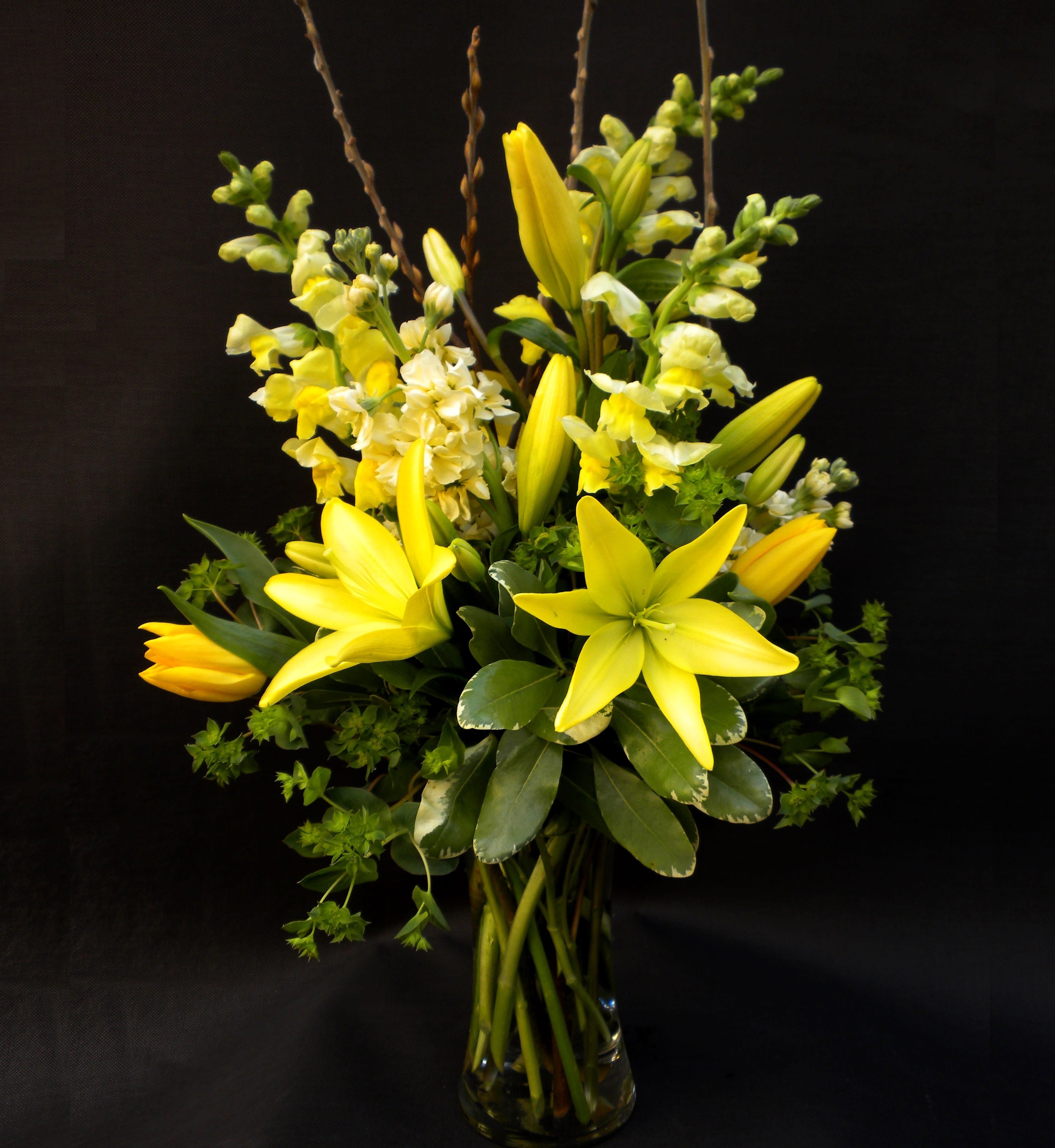 Lemon Drop - "Lemon Drop" is a radiant arrangement of cheerful yellow lilies, tulips, and snapdragons, accented with lush greenery in a clear vase. Bright and uplifting, it brings a burst of sunshine and joy to any space.