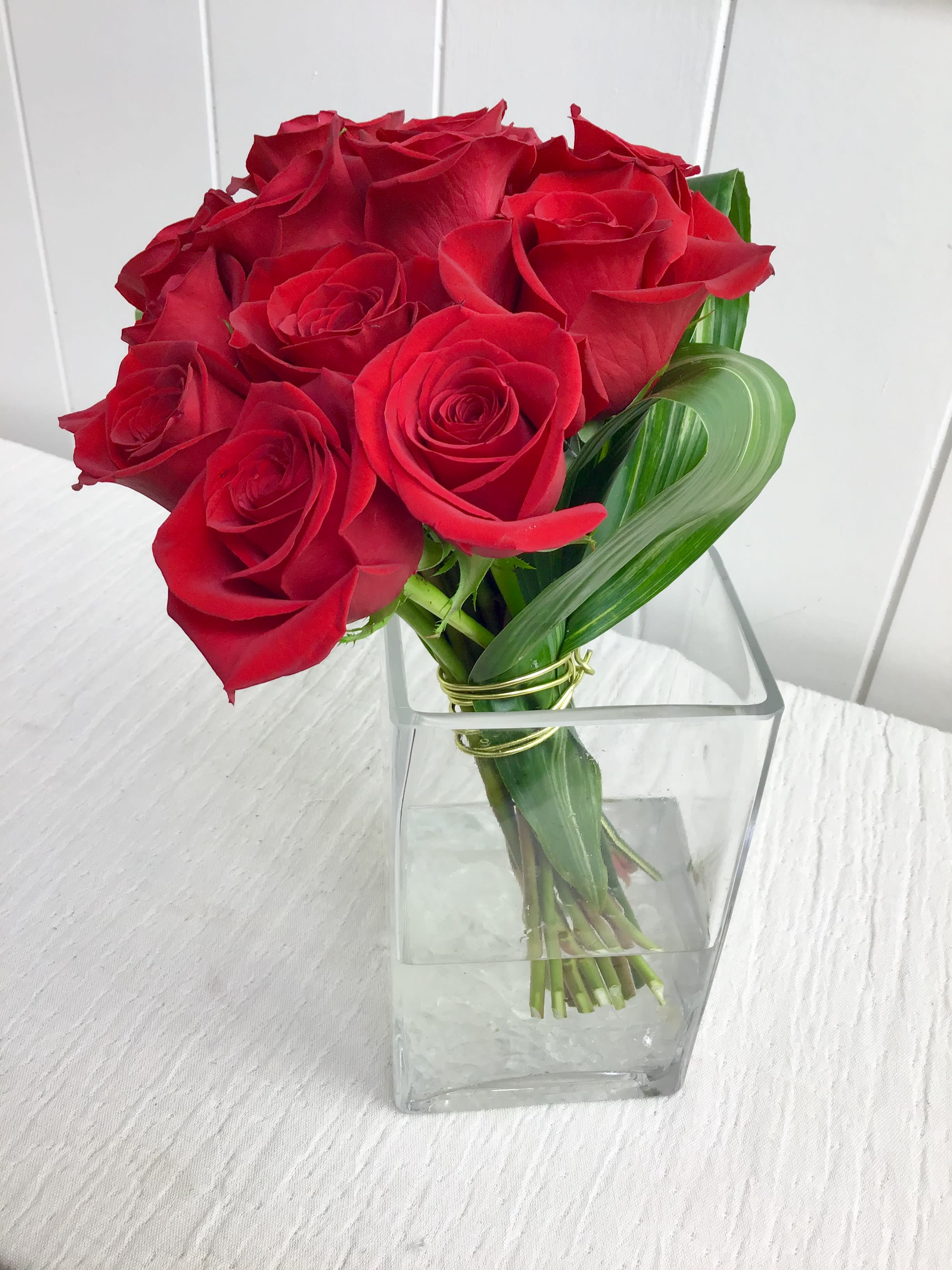 Colette - Twelve red roses are arranged in a contemporary glass vase, with diamond wire, aspidistra, and marbles. The deluxe arrangement would include fifteen roses and the premium eighteen roses. 