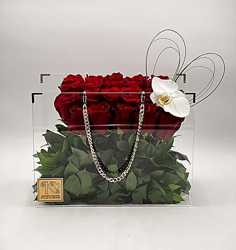 Luxe Handbag-Bold Romance - This striking arrangement features a luxurious row of premium red roses elegantly displayed in a modern clear acrylic handbag-style box. The transparent design highlights the lush green foliage beneath the roses, while a delicate white orchid accent and silver chain detail add a chic, fashion-inspired touch. Bold yet sophisticated, this design blends romance with contemporary glamour. Perfect for making a statement on Valentine's Day, International Women's Day, Mother's Day, or any unforgettable occasion.