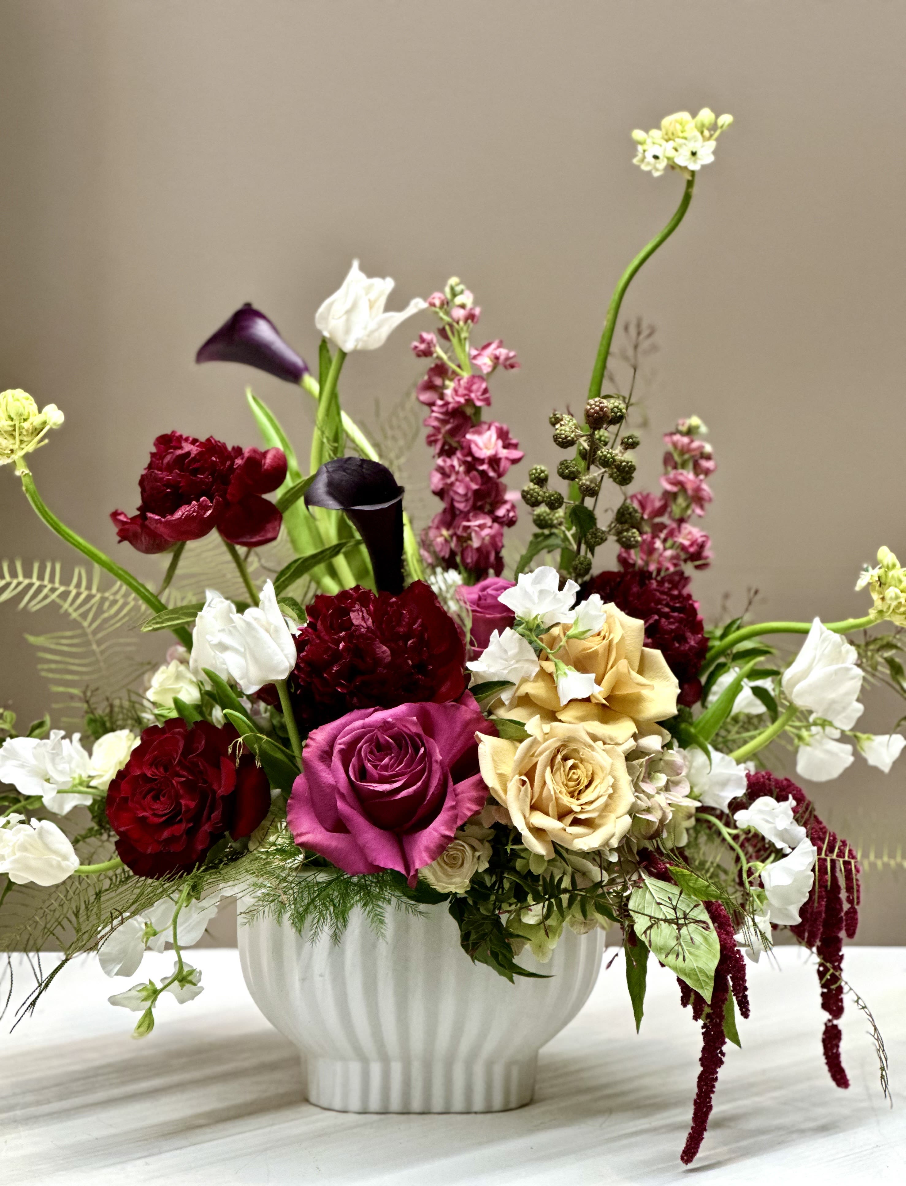 Floral Fantasia - A wonderful, carefree celebration of color and texture, this arrangement bursts with rich Burgundy Peonies, Heart Roses, and Magenta Roses, softened by Caramel-Gold Roses and delicate White Tulips. Wispy White Sweet Peas mingle with Mauve Stock and Ornithogalum, while hanging Red Amaranthus drips with drama. Deep Aubergine Calla Lilies anchor the palette, creating an arrangement that feels both whimsical and opulent.