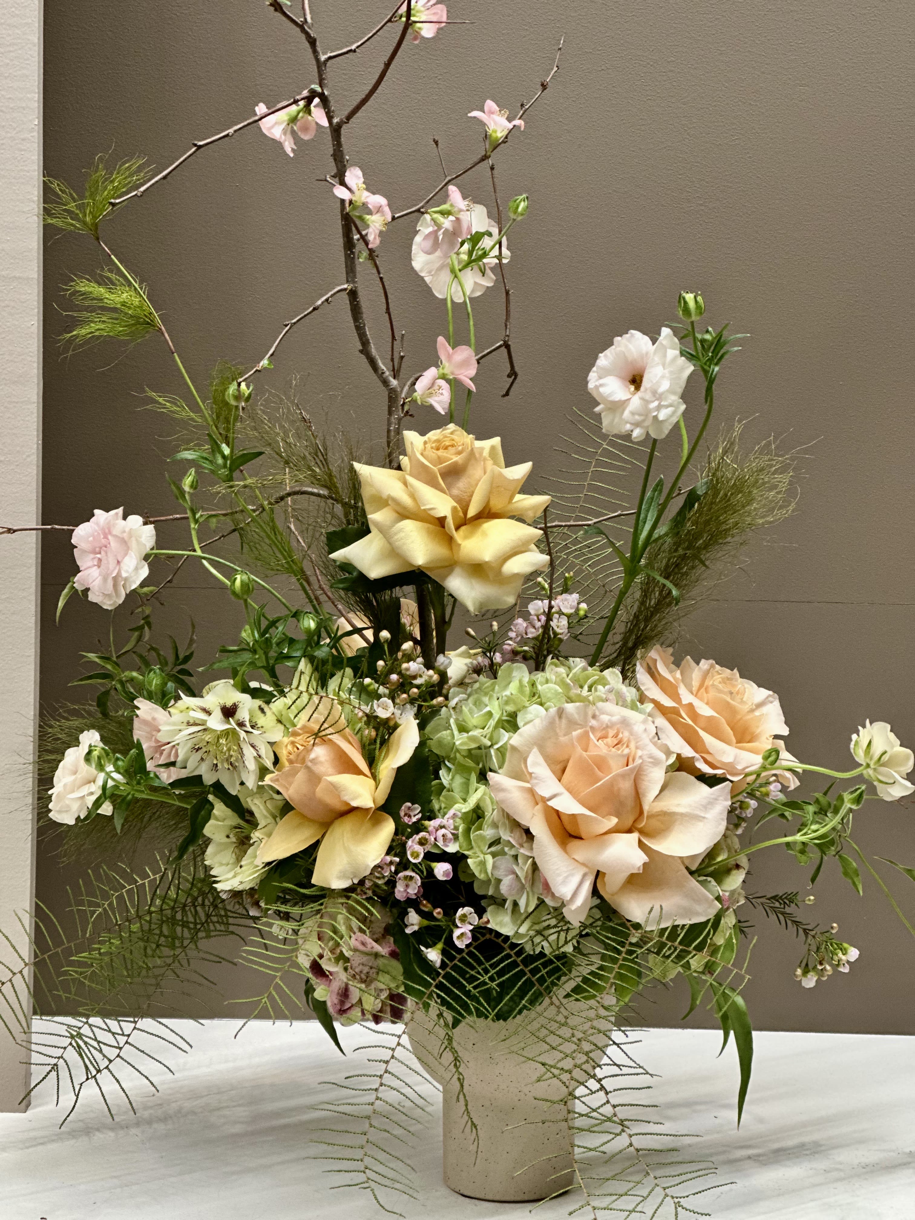 Golden Hour - A sophisticated modern arrangement in warm Gold and Peach tones featuring dutched Roses in soft caramel-Gold and pale Peach, pale Blush Waxflower, and pale Blush Butterfly Ranunculus. Hellebores and Hydrangea add subtle texture, while taller airy elements rise above the composition. Perfectly balanced in form and color, this elegant arrangement brings contemporary flair and luxury to any space.