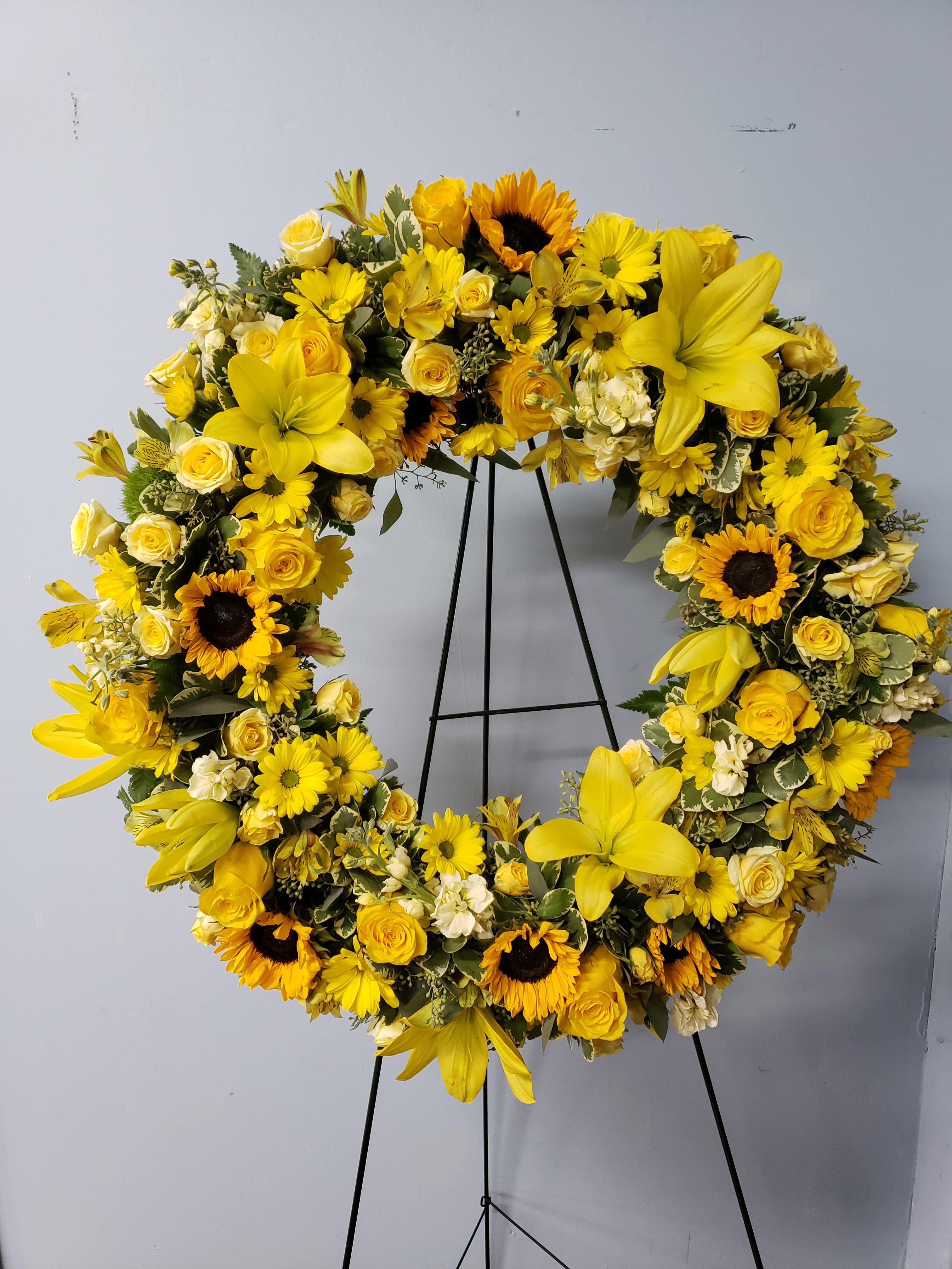 Yellow Tribute Wreath - Bright and full of life, this 18-inch wreath is beautifully adorned with the season’s finest yellow blooms. Radiating warmth and positivity, the vibrant florals symbolize remembrance, hope, and the celebration of a life well lived. Perfect as a tribute for memorial services or to bring comfort and light during times of loss.  Please note: Floral variety may vary based on seasonal availability, ensuring each wreath is fresh and unique.