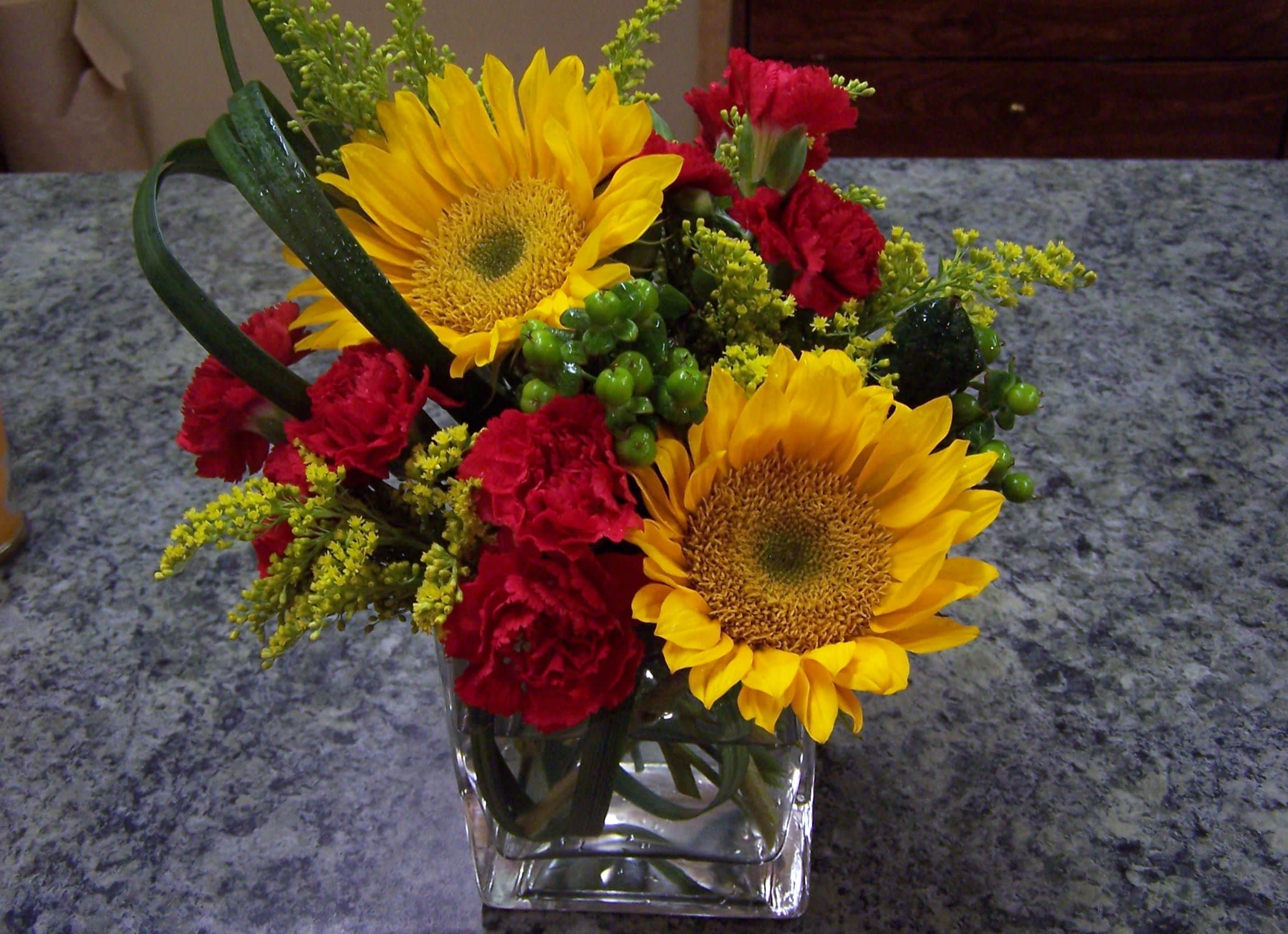 A Pick-Me-Up - The perfect gift to brighten someone's day. This 5x5 cube is filled with dancing sunflowers, hypercum berries, solidago and mini carnations with bear grass as an accent.
