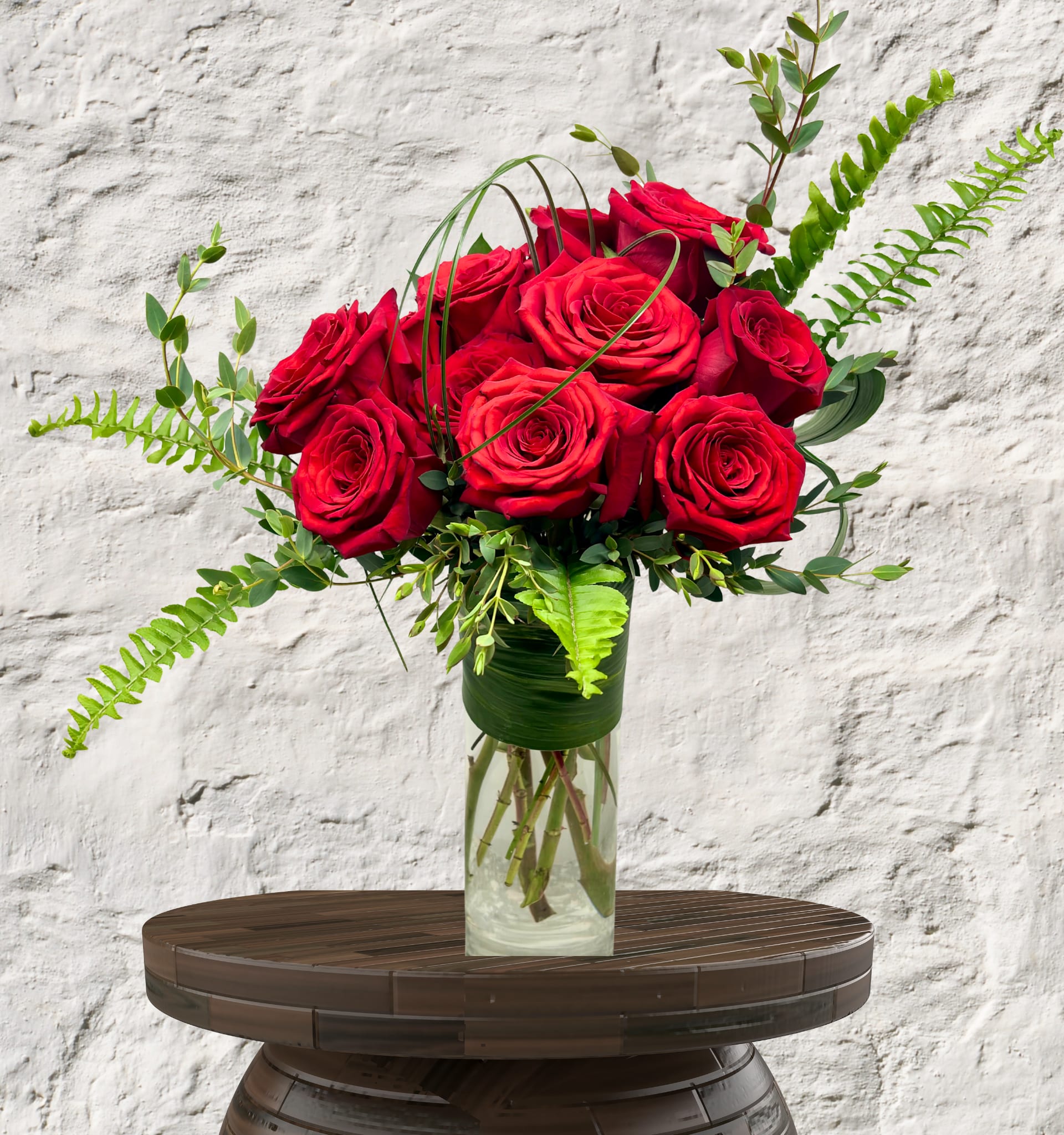 Deeply Devoted - A Modern Twist on a Timeless Tradition  Make a statement this Valentine’s Day with an arrangement that speaks the language of luxury. Our Deeply Devoted arrangement features a lush gathering of premium, long-stemmed red roses, known for their deep crimson hue and velvety petals.  This isn't your average bouquet. We’ve styled these blooms with textured Sword Ferns and delicate Eucalyptus to create a wild yet refined silhouette. To complete the look, the stems are encased in a sleek glass cylinder vase lined with a contemporary Aspidistra leaf wrap, hiding the stems and putting the focus entirely on the artistry of the flowers.  Perfect For: The partner who appreciates modern design.  An elegant centerpiece for a romantic dinner.  Expressing deep, unwavering love.  Details:  Standard -  12 premium red roses. Deluxe - 15 premium red roses Premium - 18 premium red roses  Assorted designer greenery (Fern, Eucalyptus, and Grass accents).  Signature leaf-lined glass vase included.