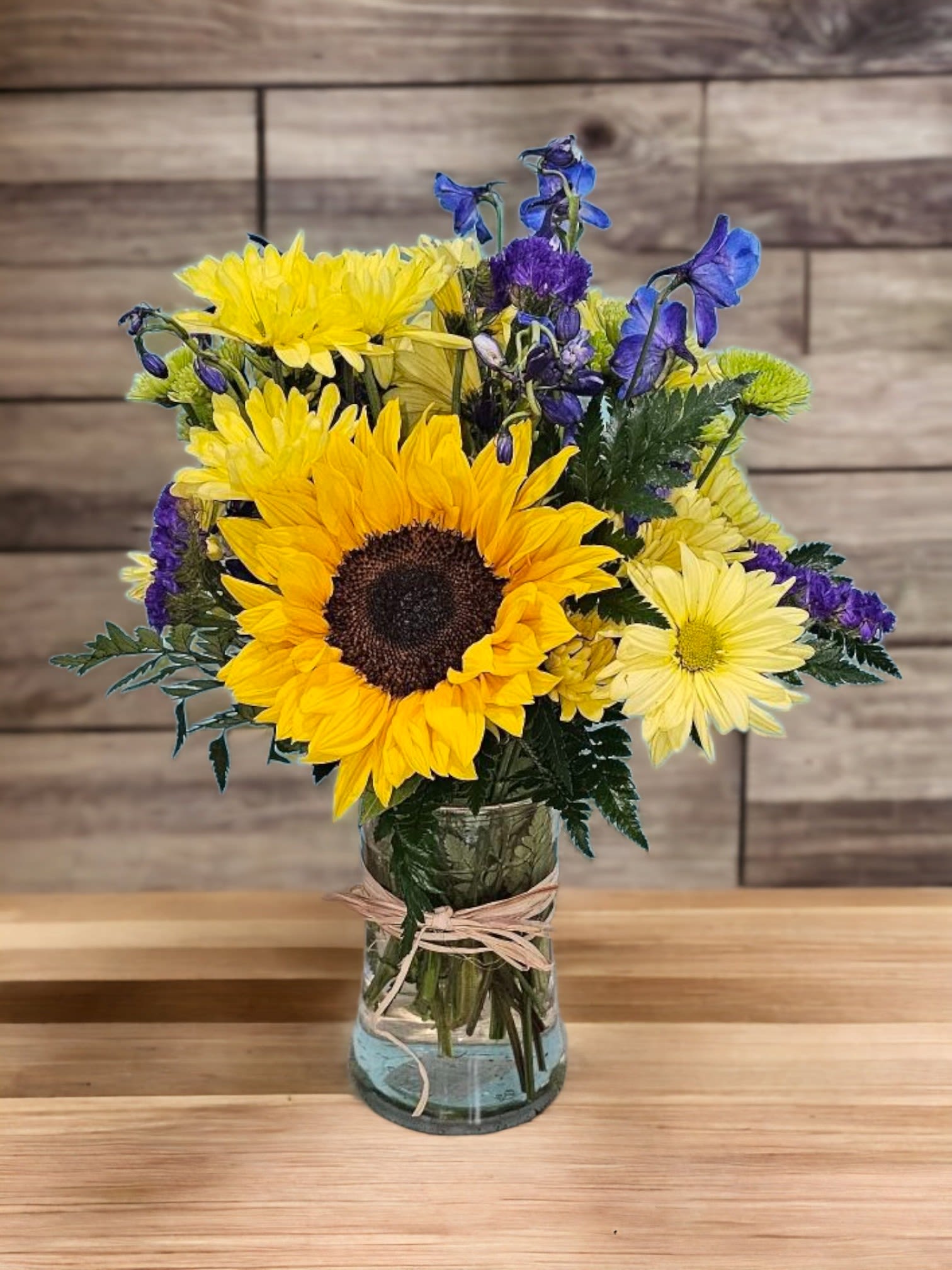 Fields of Joy Harmony - This arrangement evokes imagery of sunny, vibrant landscapes with a mix of cheerful flowers and greenery, like Sunflower, Blue Delphinium, Green Botton, some filler and fine Greenery. They capture the essence of brightness, harmony, and natural beauty that these flowers bring to a floral arrangements