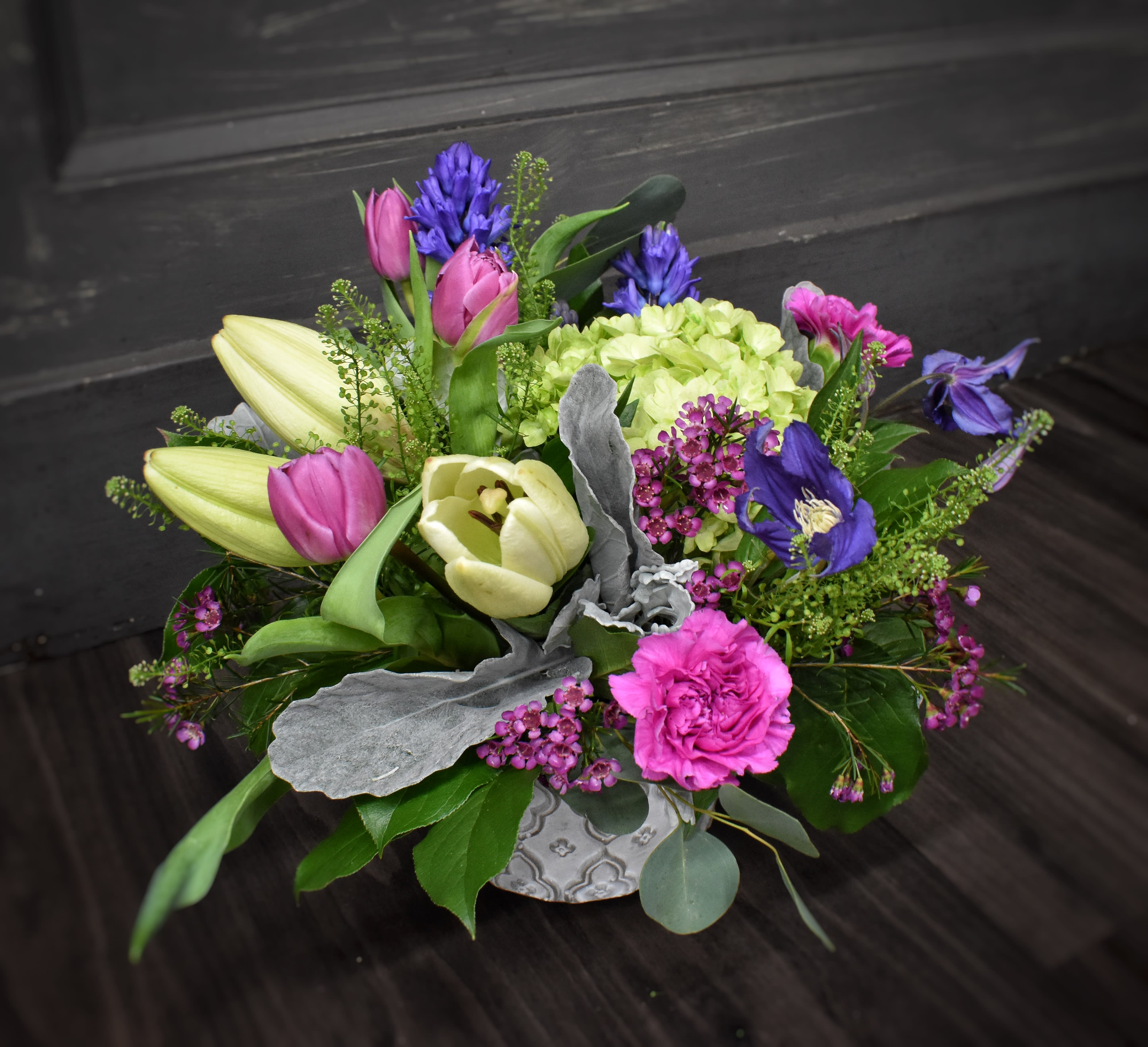 Garden Gathering - An embossed ceramic container filled with cream and shades of purple flowers. Fragrant hyacinth, lily, hydrangea and tulips fill out this beautiful floral gift. appx 12"h x 10"w