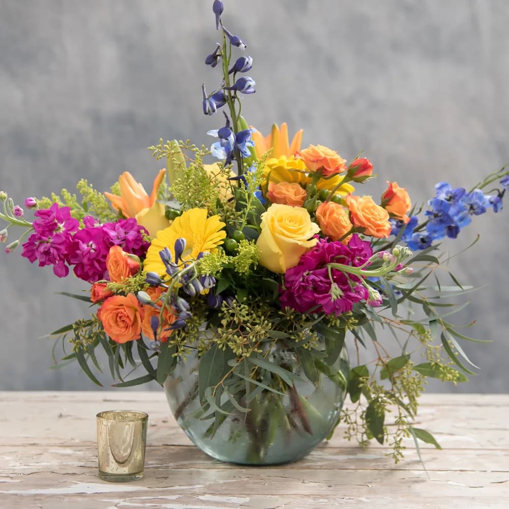 Fiesta - Let this bursting with color arrangement be the start to your party, whether it's a birthday, anniversary or just because - this bubble bowl is overflowing with orange lillies, yellow gerber daisies, fuchsia stock, blue delphinium and much more including standard and spray roses. It's Fiesta time!  