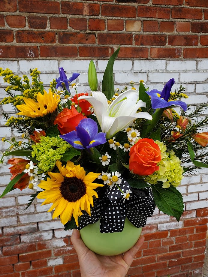 Good Cheer Grand - Bursting with color and joy, this radiant flower arrangement is a true celebration of sunshine and happiness. At its center are bold, golden sunflowers, their cheerful faces and large, textured petals capturing the warmth of a summer day. 