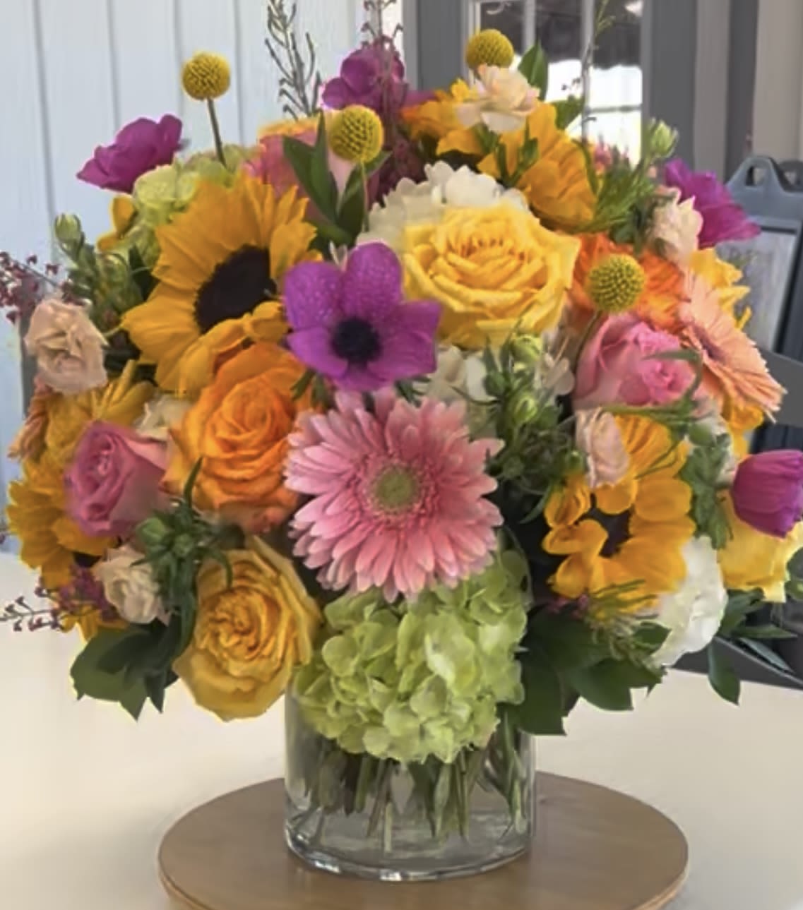 Sun-Kissed Garden Bliss - A radiant, cheerful arrangement bursting with color and charm. Featuring vibrant sunflowers, pink, golden and coral roses, playful pink and peach gerberas, delicate anemones, and lush green and white hydrangeas in a clear glass cylinder. 