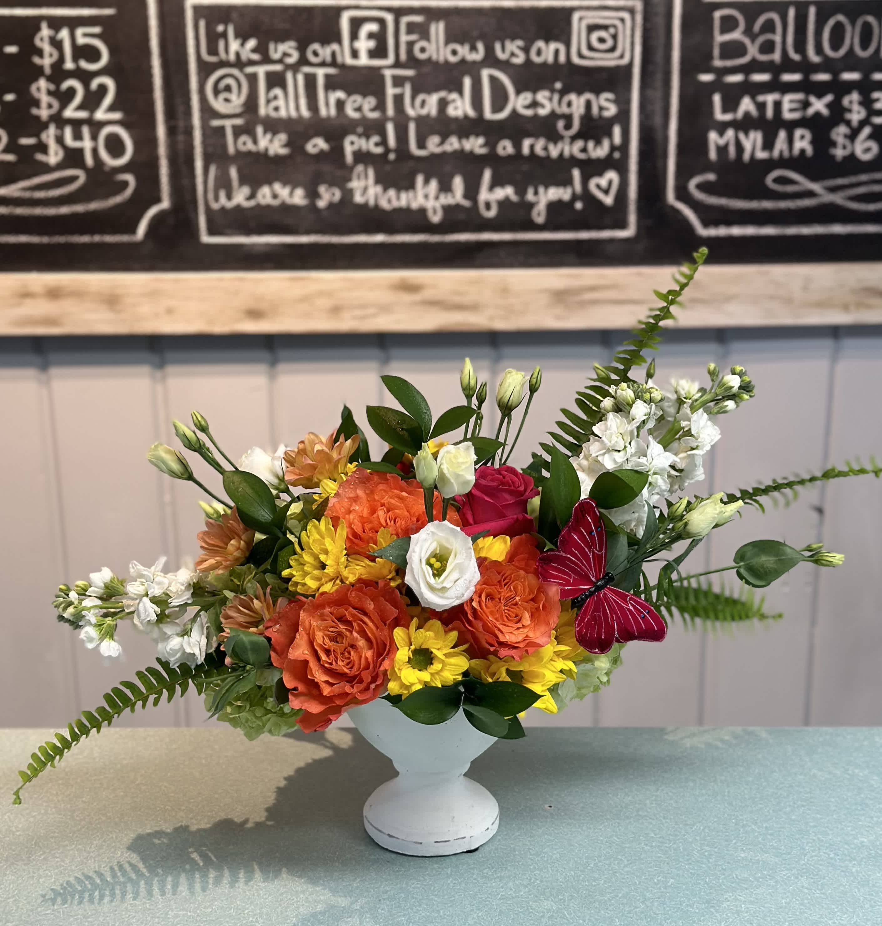 Sunburst - Free spirit roses, hot spot pink roses, white lisianthus, yellow daisies, white stock, peach daisy, sword fern with a butterfly in a small white ceramic pedestal