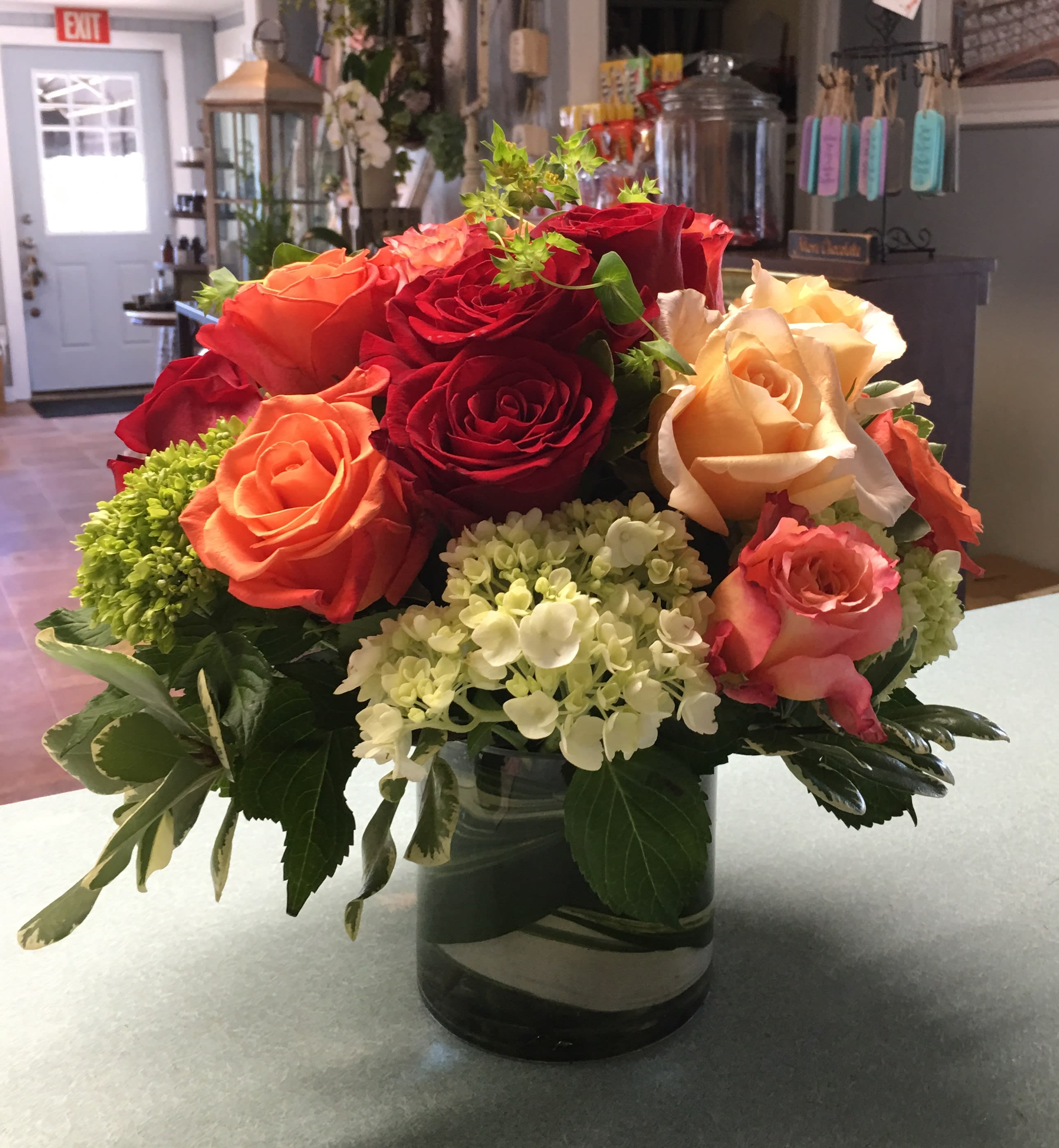 Medley of Orange and Red Roses with Apple Green Hydrangea - Crisp red and orange colors adorn these roses that mimic the beautiful foliage of the Fall season.  A hint of apple green hydrangea offer the perfect backdrop to these lush colors. 5 inch cylander with tropical foliage lining the inside of the vase.  