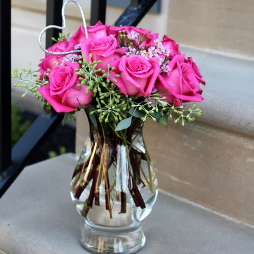 Basic Beauty - "Our ""Basic Beauty"" arrangement is perfect for enhancing the feeling and decor in any household, business, or event. Looking for a simple yet sophisticated design? Select the ""Basic Beauty"" arrangement!  Included are a dozen pink roses, embellished by stems of seeded eucalyptus and cute pink wax flowers. The clear vase helps support the pop of color, which can be adorned with a matching ribbon of your choice. Accessories included: One heart pick."