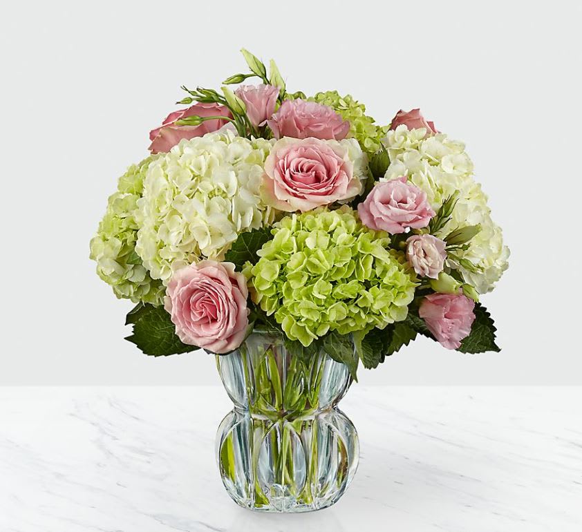 Always Smile™ Luxury Bouquet - VASE INCLUDED - The play of pink against green and white can't help to bring a mirthful beauty to this stunning bouquet. The blushing swirl of the roses and lisianthus bring a sweet drama to this arrangement offset by clusters of green and cream hydrangea. With the vase tinted in a subtle blue shade, to add even further interest, sparkling as it catches the light through each facet of it's unique design, this gorgeous flower arrangement is ready to set the stage for a treasured moment of pure happiness. IMPRESSIVE bouquet includes 11 stems. Approx. 15"H x 15"W. STUNNING bouquet includes 19 stems. Approx. 19"H x 18"W