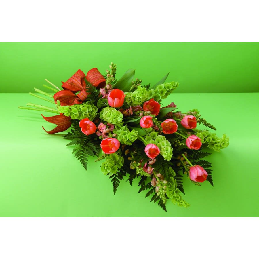 Bells of Comfort - This bright casket piece offers honest sympathy and tenderness with a selection of blooming tulips, complemented by bells of Ireland and an adorning red ribbon. Your loved one will be comforted in spirit. 