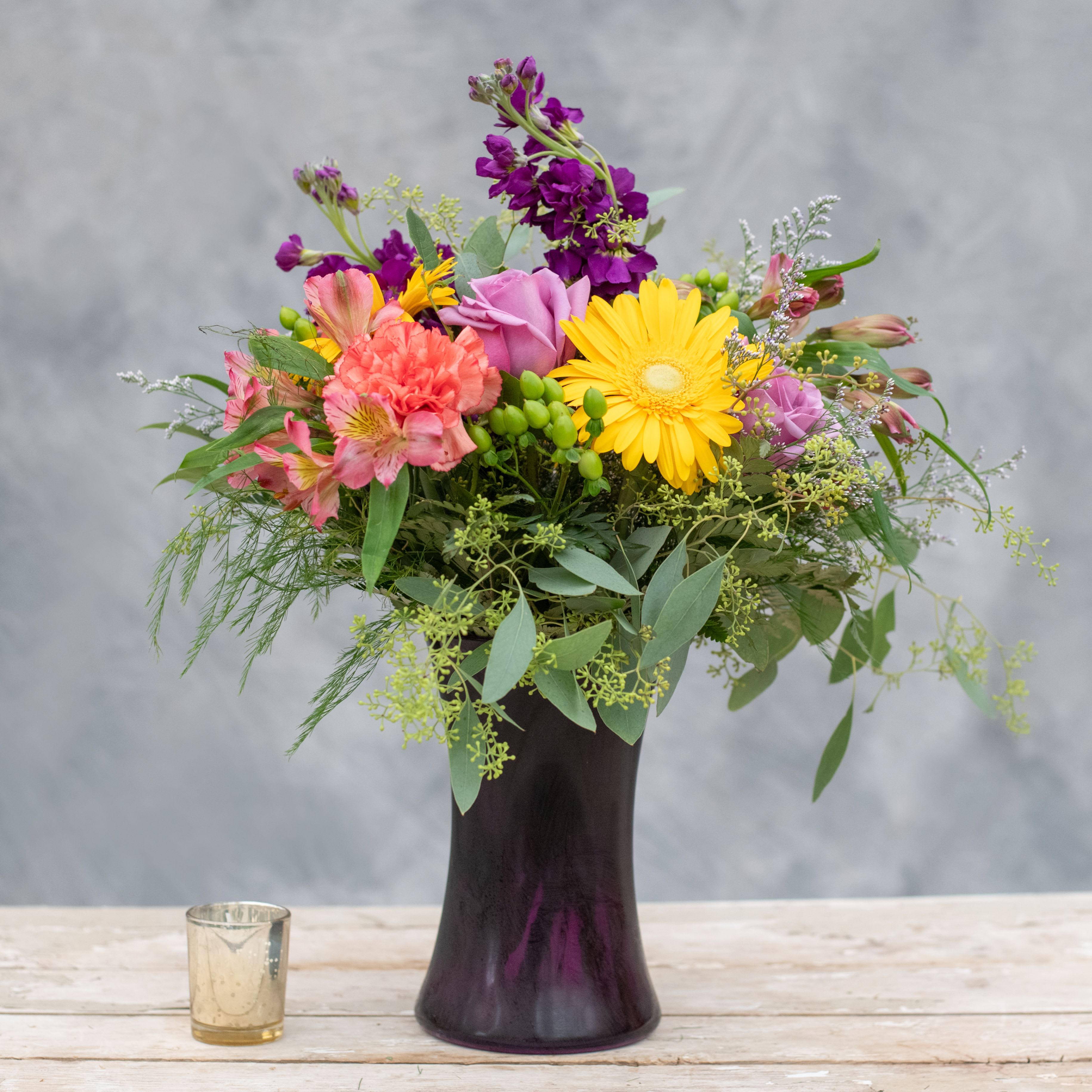 Mardi Gras - Get in the festive spirit with this jewel tone arrangement in a purple glass gathering vase.  It's filled with purple stock, yellow gerbera daisies, orange carnations, lavender roses and more.  