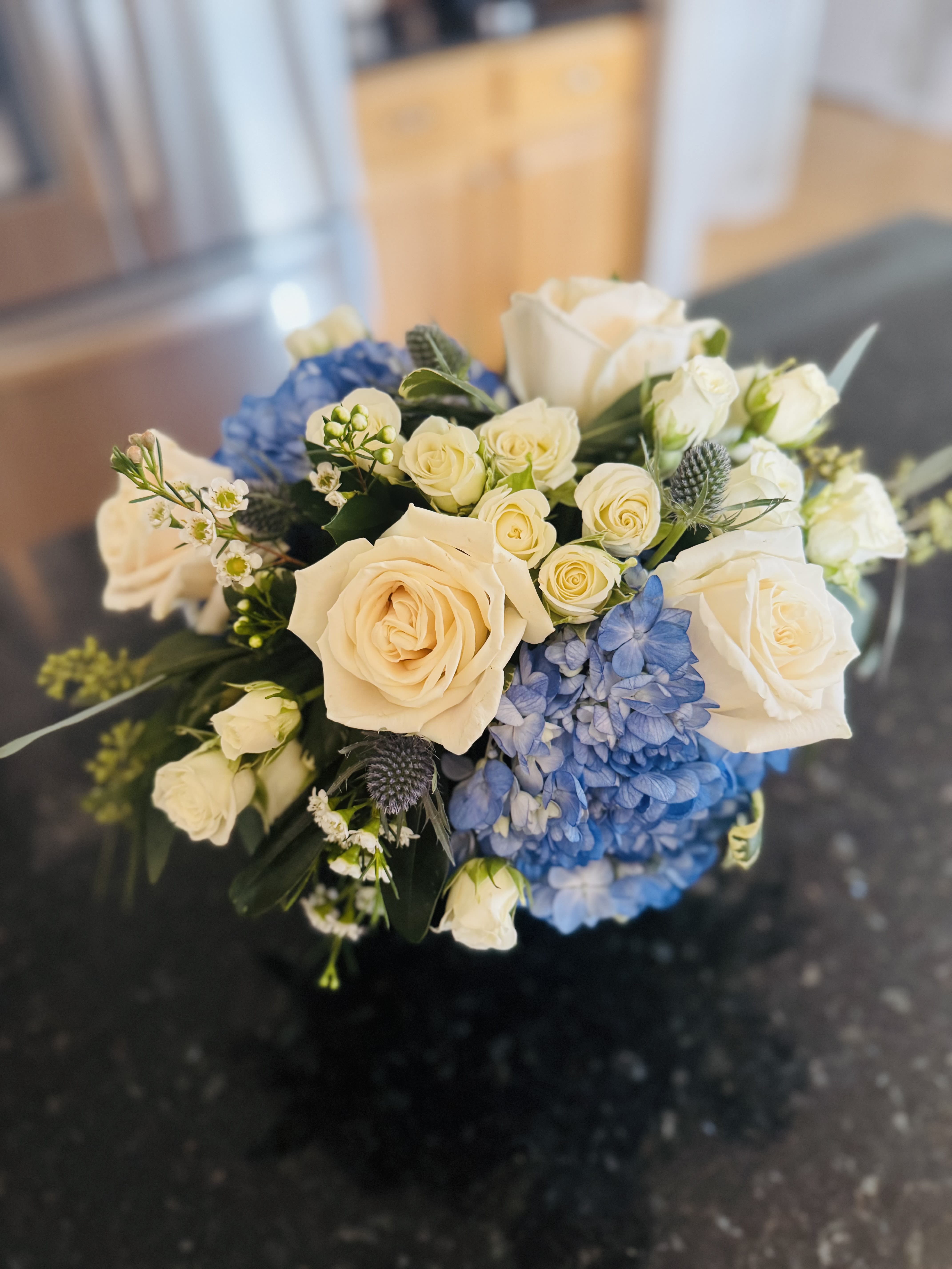 Blue Bayou - blue Hydrangea, white Roses, wax flower,blue thistle arranged in cylinder.