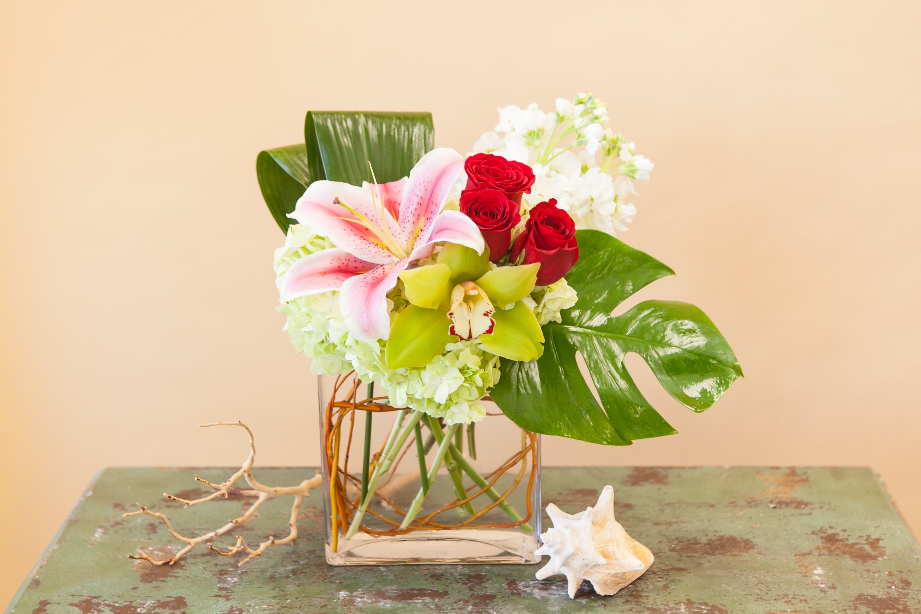 Modern Tropics - A beautiful and contemporary combination of red roses, hydrangea, lilies, cymbidum orchids and fragrant stock with tropical leaves. Flower colors may vary from week to week, please call if this is a concern.