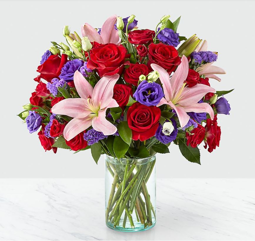 Truly Stunnin Bouquet - Share a smile with your loved ones through a bouquet filled with stunning beauty and heartfelt joy. Comprised of vivid red roses, purple double lisianthus, pink lilies and red spray roses within a clear glass vase, vibrant color bursts from every bloom. Make their day brighter with a gift that is Truly Stunning™!