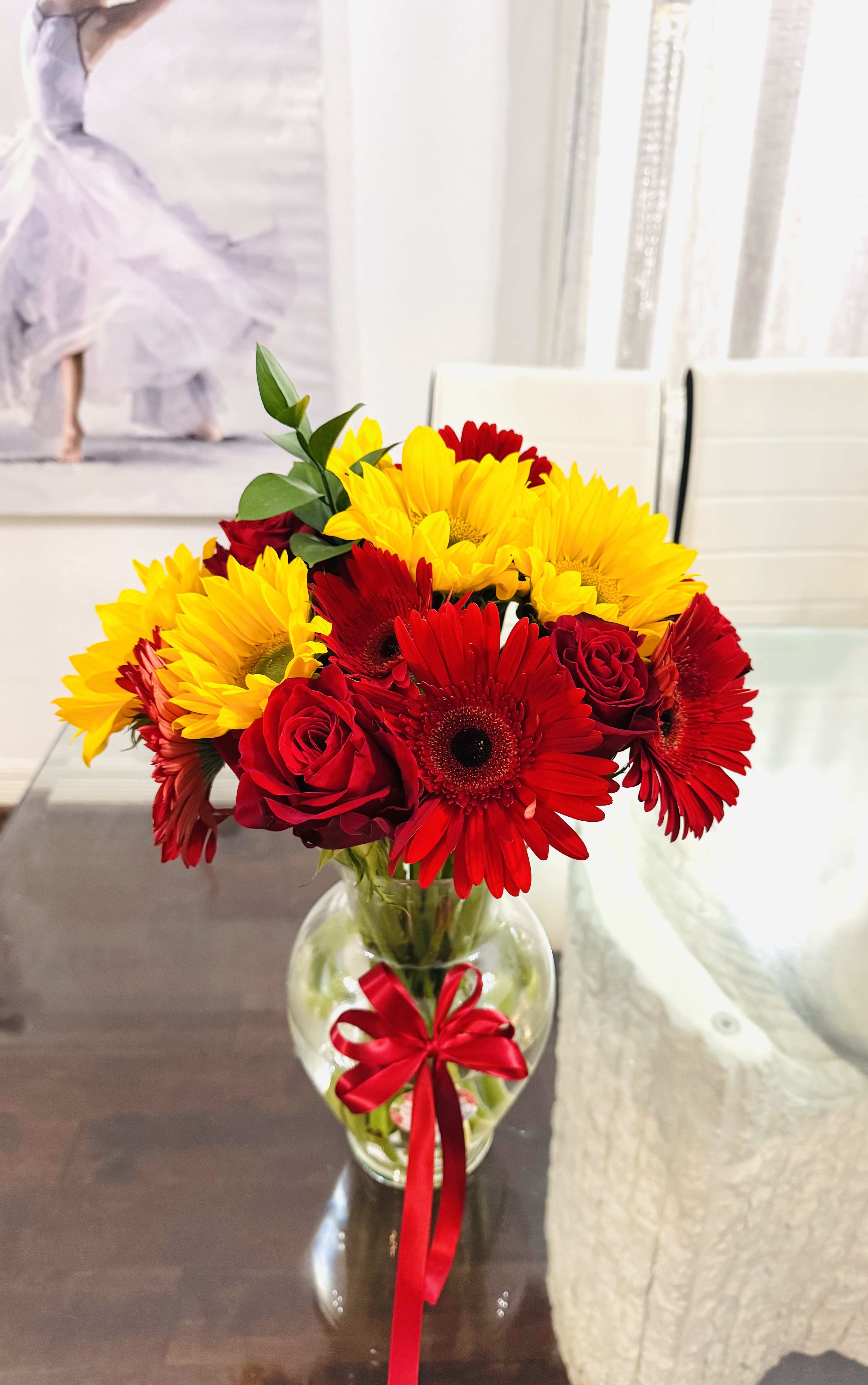 Joy and Grace Gerbera with roses - This Bouquet is a striking composition of romantic fire and golden light, blending the timeless allure of red and pink roses with the radiant joy of sunflowers, and Gerbera. Each bloom plays its part in a beautiful contrast- the roses deep crimson, speak of devotion, desire, and enduring love. The sunflowers, bold and sunlit, bring brightness and vitality, their golden faces turned as if to follow hope itself.