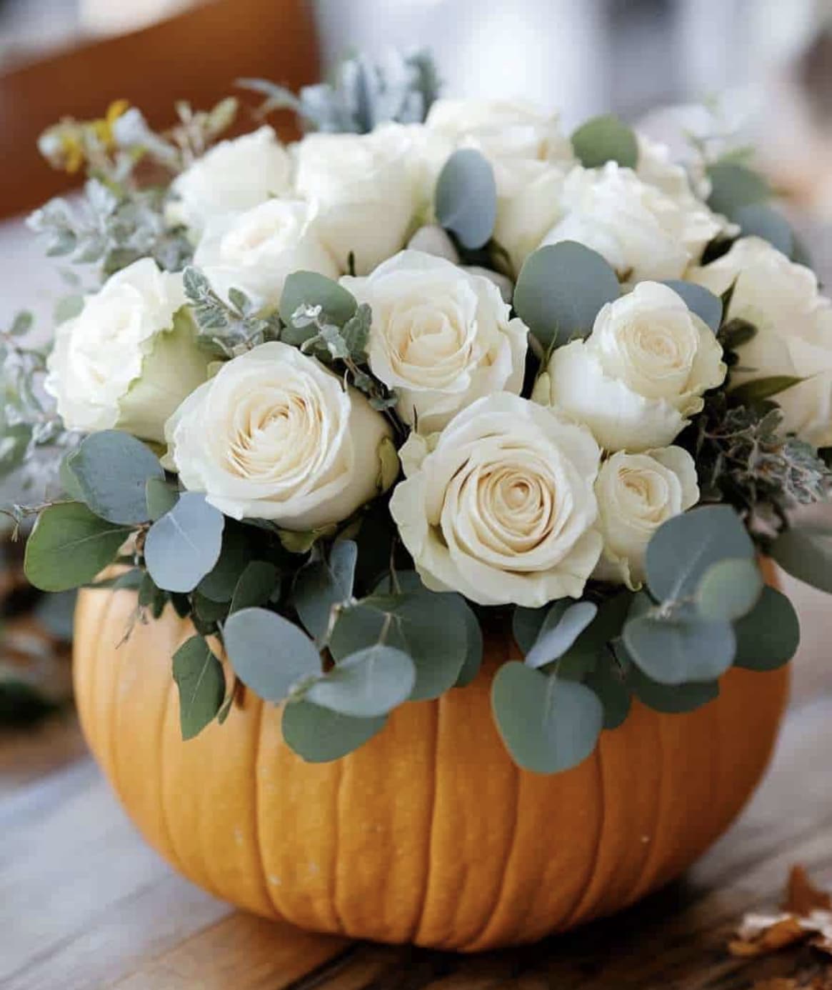 Elegant Thanksgiving - This design is sophisticated and graceful centerpiece that blends rustic harvest charm with timeless floral arrangement. A classic pumpkin vase transformed into s work of art, holding a lush bouquet of creamy white roses paired with a soft eucalyptus foliage. The ivory blooms add a touch of purity and elegance, while the muted green eucalyptus leaves provide texture balance. The warm pumpkin base contrasts beautifully with the delicate roses, creating a piece that feels both seasonal and refined.  This design is perfect for Thanksgiving gatherings, fall weddings, or upscale autumn decor- a statement piece that combines the coziness of autumn with the luxury of roses. 