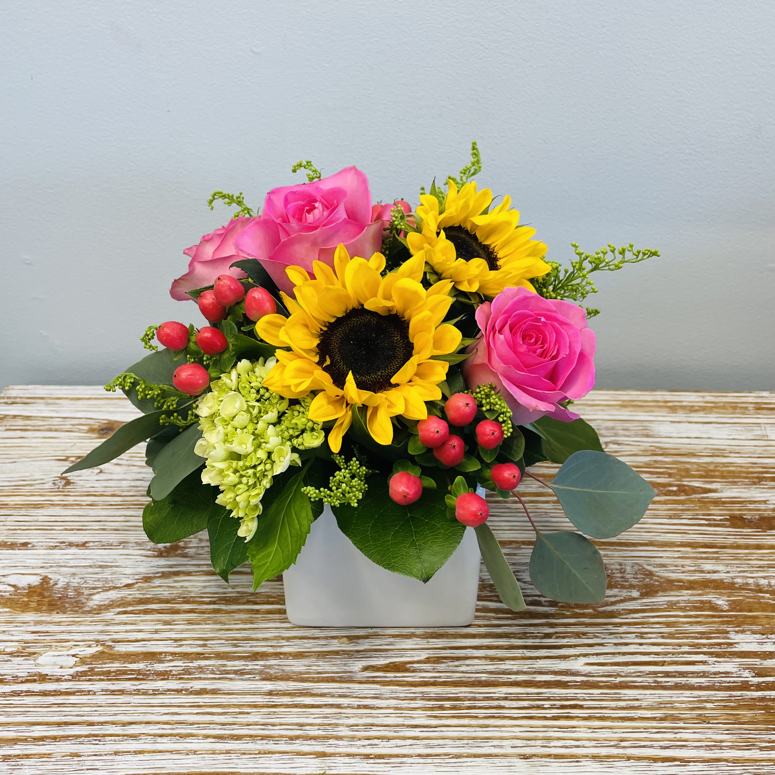 Farm Girl - A bright and beautiful arrangement consisting of Sunflowers, bright Pink Roses, Pink Berries and miniature Green Hydrangea in a 4" White Ceramic Cube.  Approximately 10” Tall  Container Country of Origin: China Flower Country of Origin: Ecuador and/or Columbia