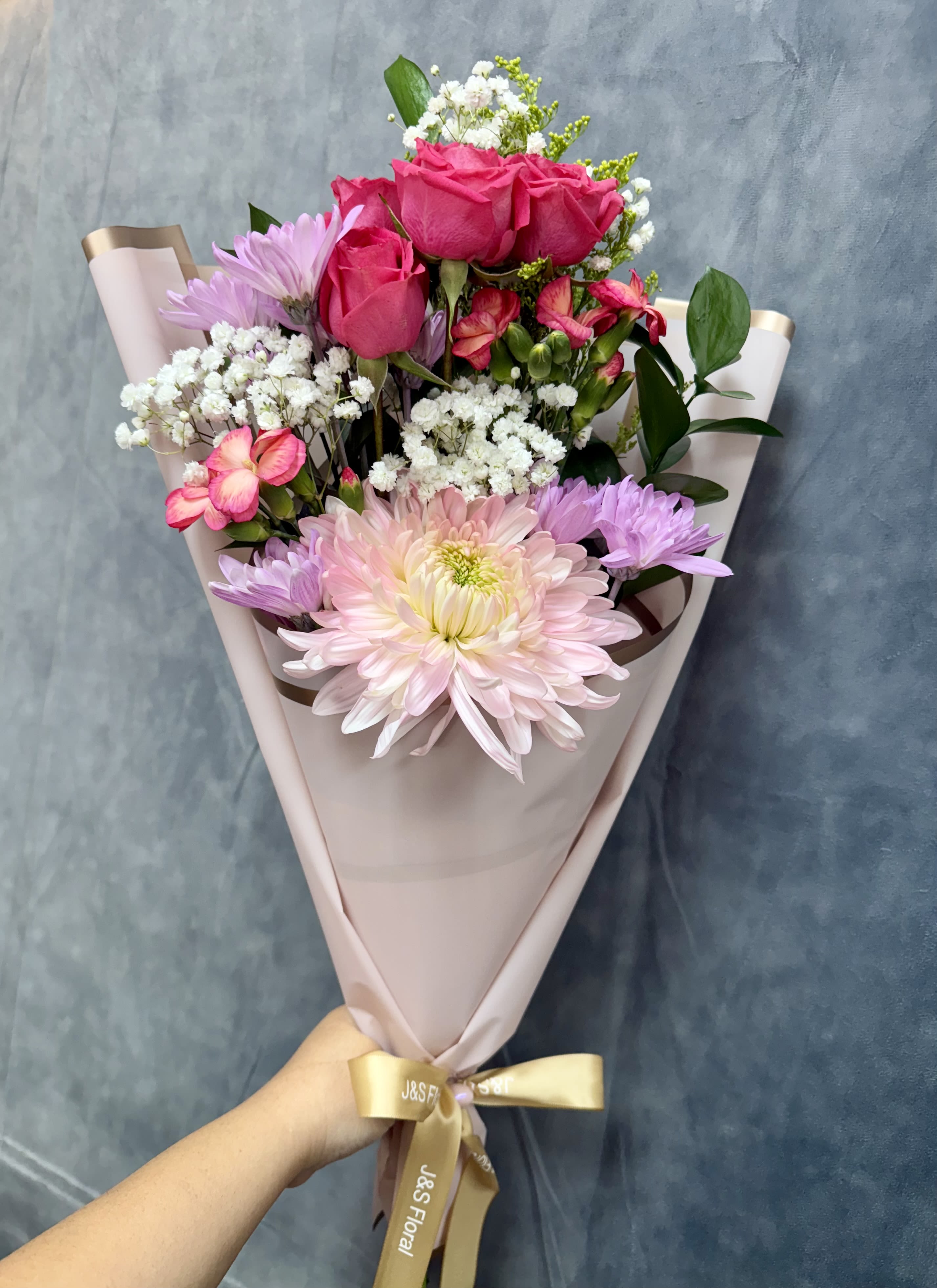 Pink Breeze Bouquet  - Our petite Pink Breeze hand-tied bouquet features cheerful mums, spray roses, daisies, and mini carnations, all wrapped in soft pink paper for a sweet, charming touch — perfect for brightening someone’s day.