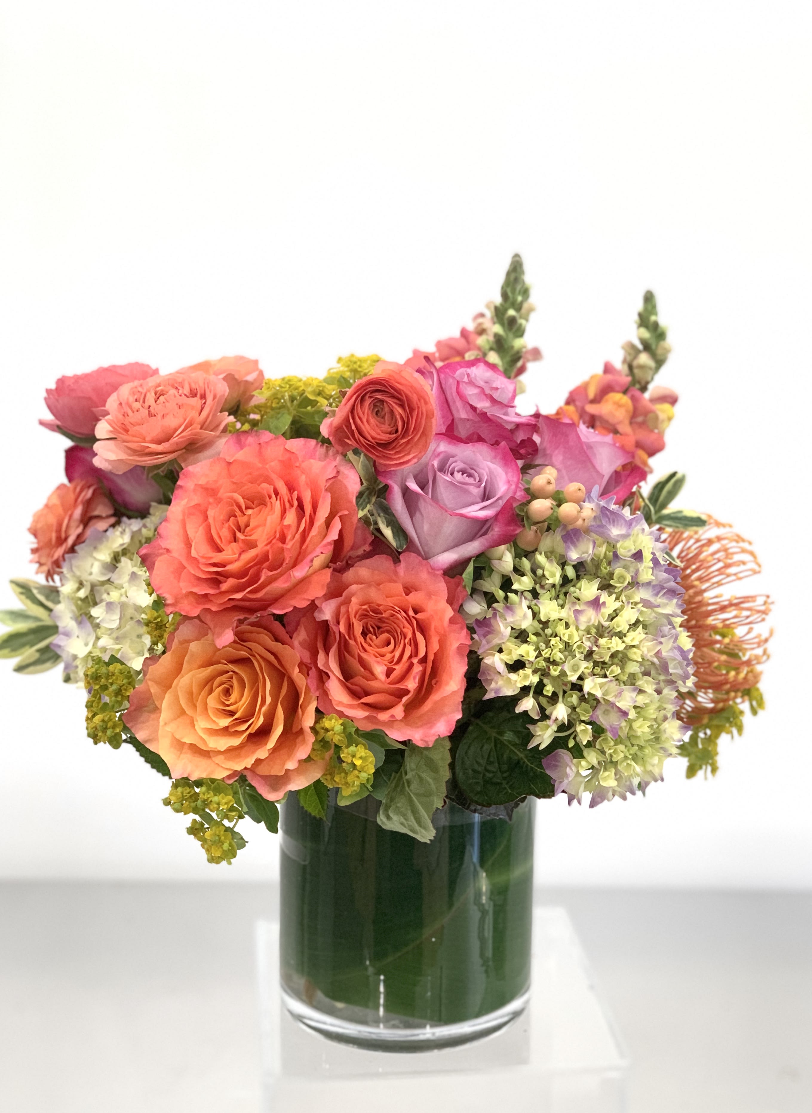 Burst of Sunshine - Lush arrangement bursting with orange and lavender florals set in a ti leaf lined vase. A burst of color for a dining room table, credenza, or coffee table. Designers choice in orange, purples, soft greens.   Saturday and Same Day are available if you call the shop.   ** 415 751 2747 10:00AM -5:00PM