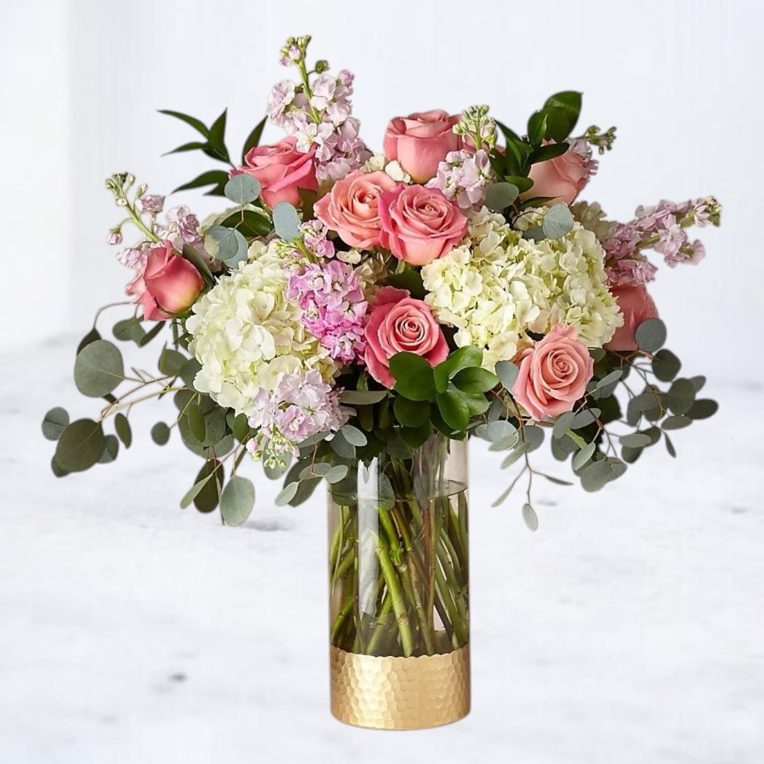 Enchanted - Step into an enchanted world with our "Enchanted" bouquet. This lush arrangement features a beautiful mix of pink roses, soft lilac blooms, and white hydrangeas, creating a delightful blend of colors and textures. Accented with delicate greenery, this bouquet is elegantly arranged in a tall glass vase with a chic gold base. Perfect for special occasions or simply bringing a touch of garden elegance into any space, this arrangement is available for delivery in Galveston Texas, and surrounding areas. Order now to experience the enchanting beauty of this floral masterpiece!