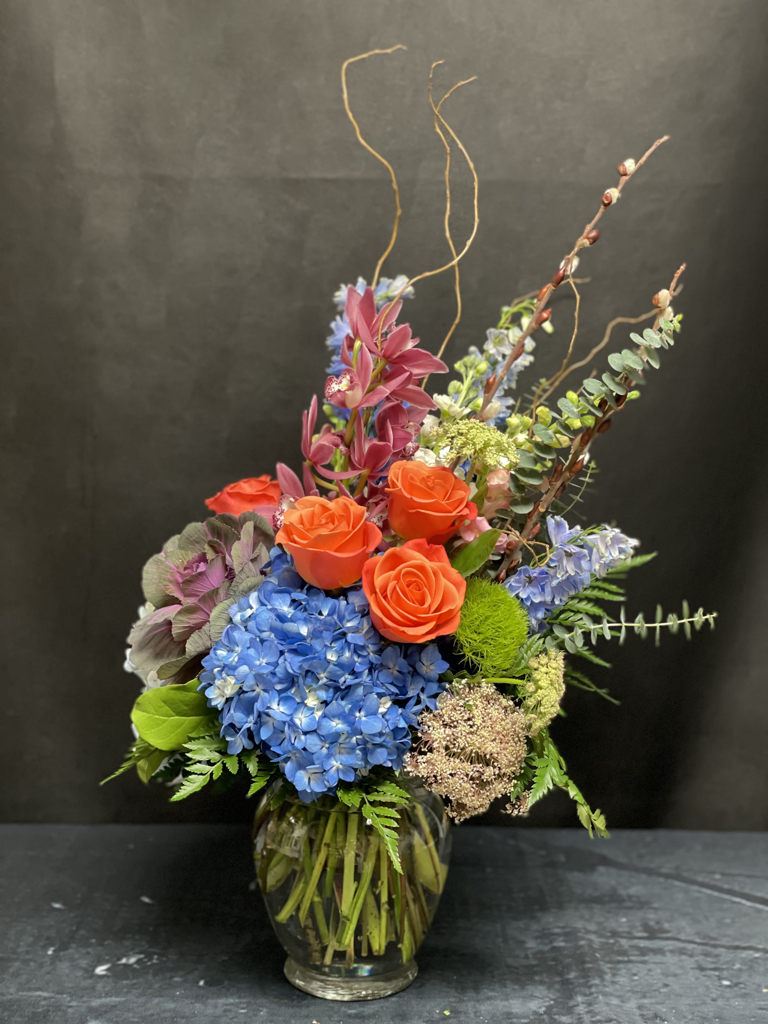 Wildest Dreams - A vibrant, whimsical dance of color and texture. Featuring lush blue hydrangeas, bold orange roses, delicate mini cymbidiums, and airy queen Anne’s lace, this arrangement is grounded by earthy purple kale, dramatic blue delphinium, fragrant stock, and sculptural curly willow. Designed to stir the imagination and leave a lasting impression — just like a dream you never want to wake from.