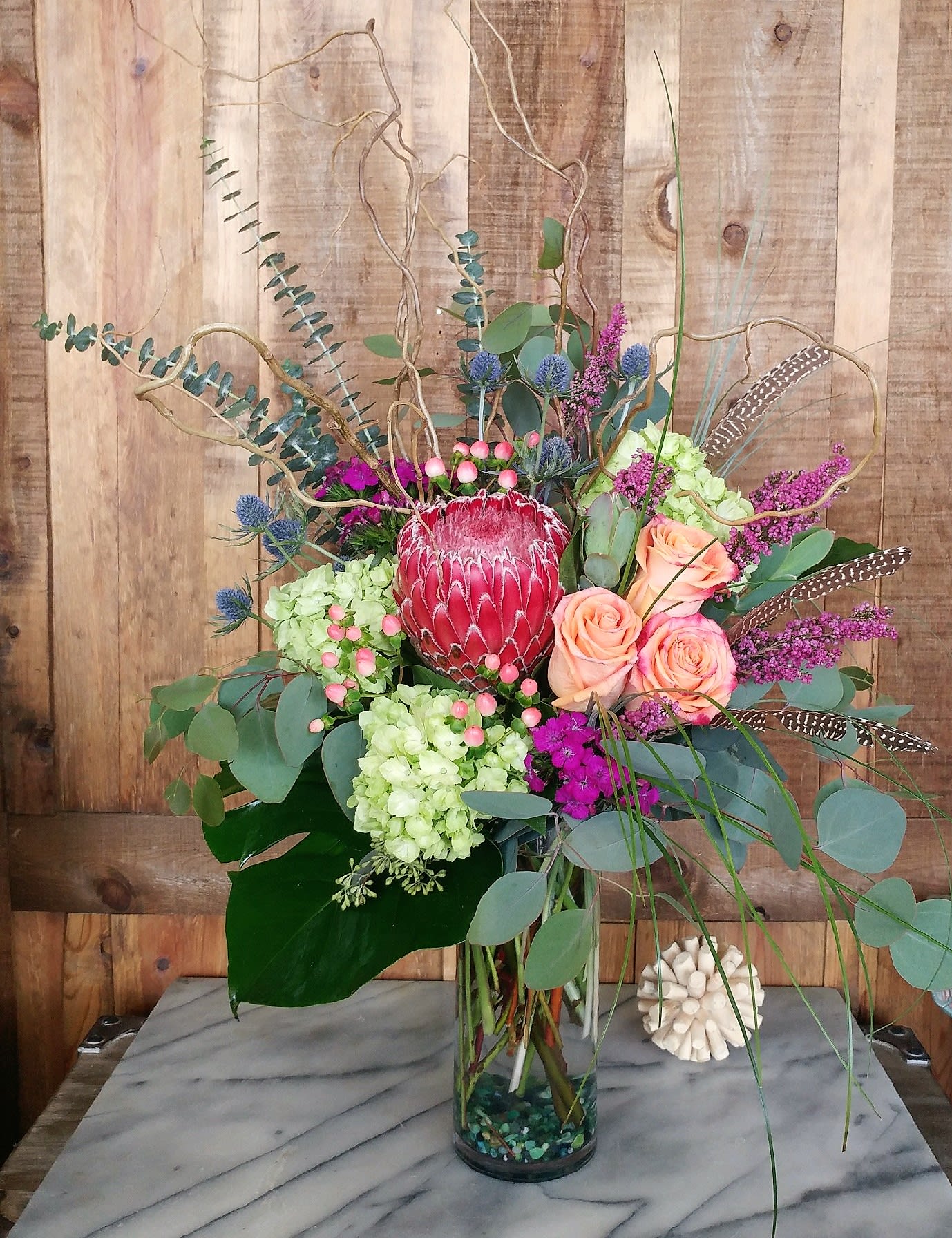 Bohemian Beauty - Bohemian Beauty – A Luxe Botanical Arrangement for the Bold &amp; Beautiful in Las Vegas  Make a powerful statement with this wild yet refined floral masterpiece, expertly designed for those who crave originality and sophistication. Anchored by a dramatic protea, this arrangement bursts with textures and colors—lush green hydrangeas, coral garden roses, hypericum berries, thistle, and wisps of eucalyptus. Curly willow branches and pheasant feathers rise organically, adding height and intrigue, while the tall, clear vase gives a modern, airy finish.  Perfect for upscale interiors, luxury gifting, or events that demand a unique floral focal point, this design captures the spirit of the desert with a touch of exotic flair—crafted fresh in Las Vegas with only the finest blooms.