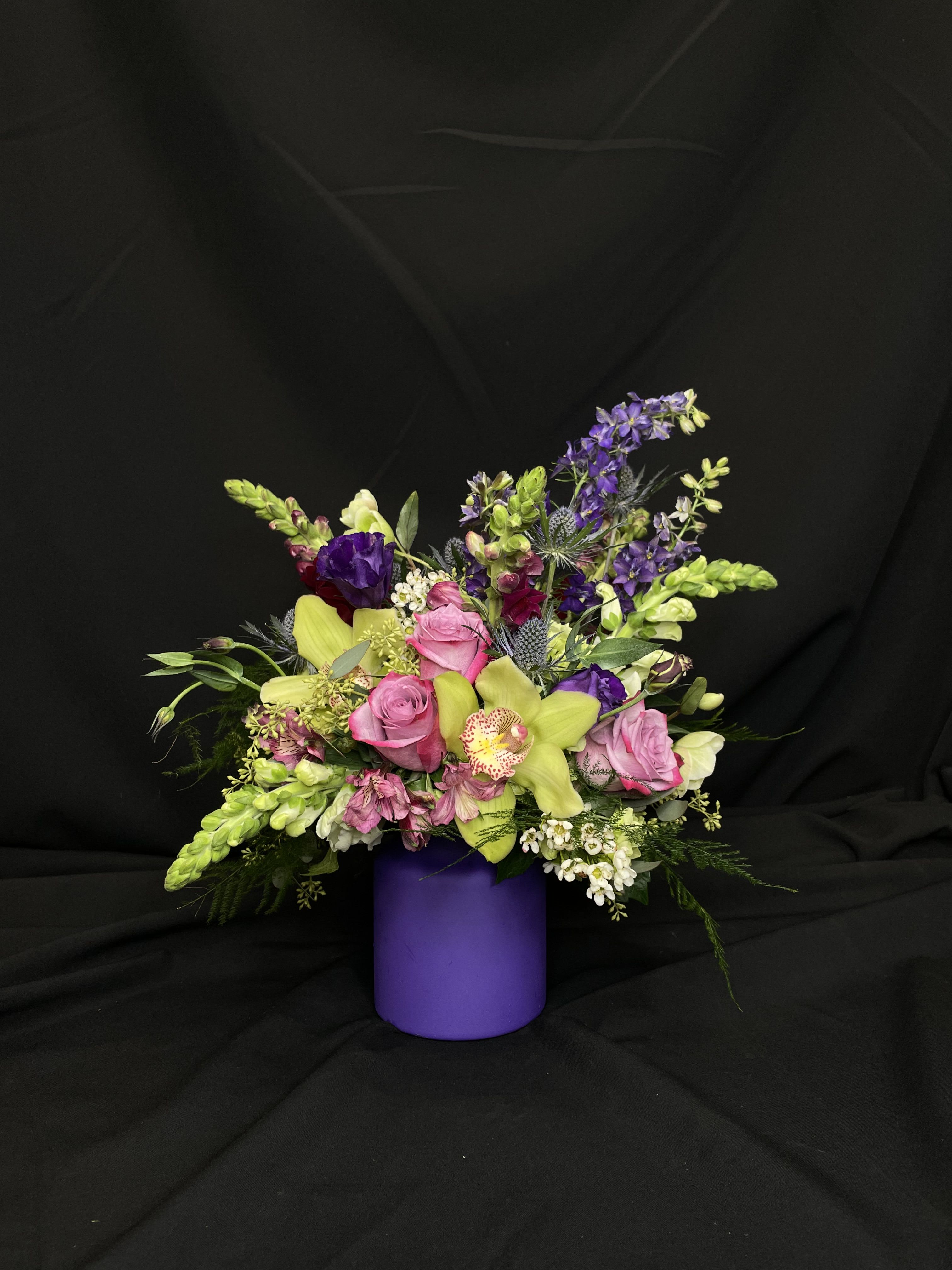 Twilight Enchantment - Dive into a dreamscape of cymbidium orchids, larkspur, roses, snapdragons, eryngium, alstroemeria, lisianthus, and lush greenery in a purple cylinder vase. The color palette includes deep purple, lavender, chartreuse, white, and burgundy blooms. Perfect for making a bold statement or celebrating someone who appreciates the extraordinary.