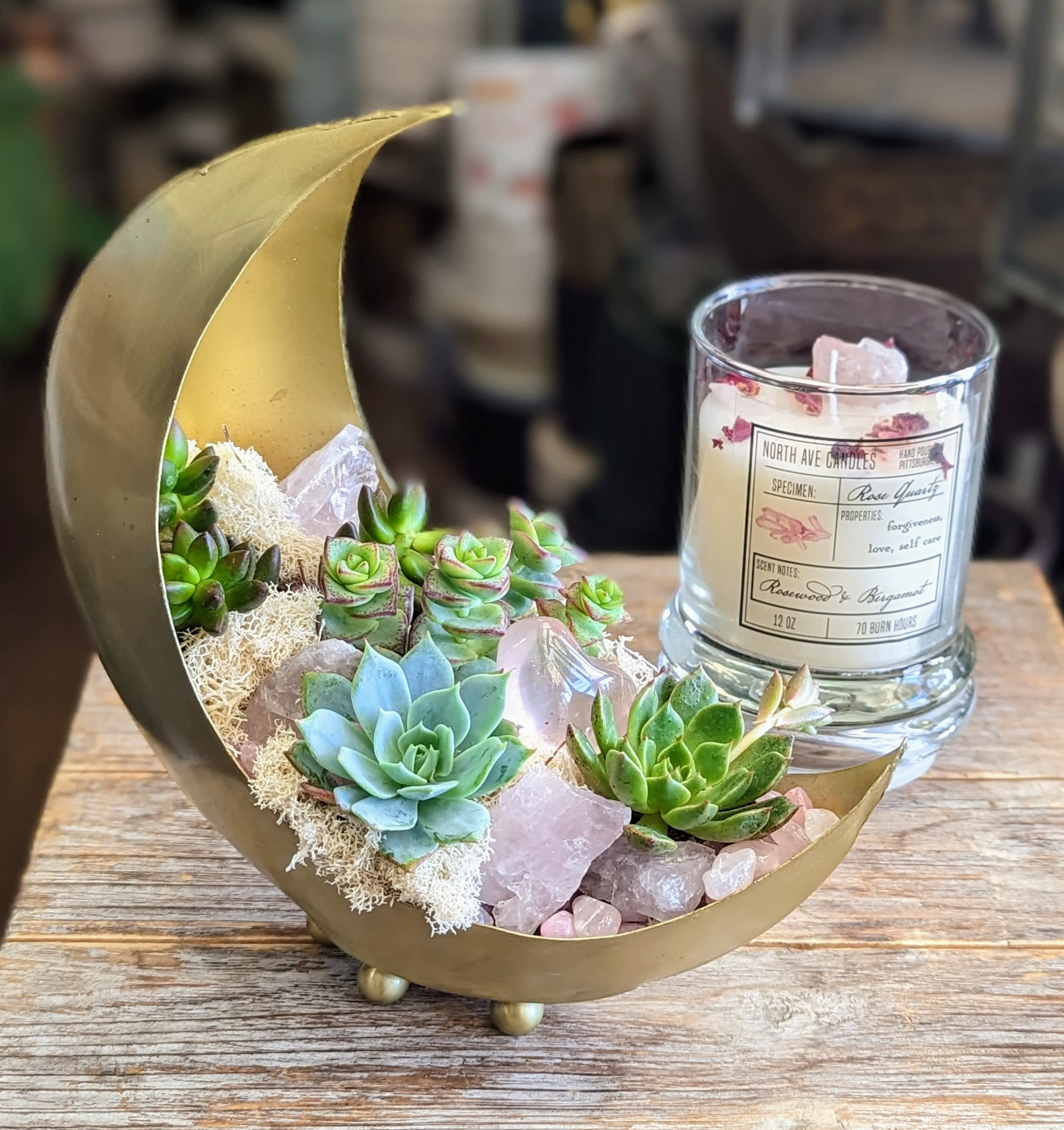 Mini Luna And Rose Quartz- Rosewood And Bergamot Candle- 12oz - Delicate, dreamy, and full of cosmic charm — Mini Luna is a petite celestial garden designed to enchant. Cradled in a golden crescent moon vessel, this miniature succulent arrangement features an array of lush rosettes, soft moss, and glistening rose quartz crystals. Thoughtfully styled with intention and heart, Mini Luna radiates serenity, love, and grounding energy.  A perfect gift or accent piece from Windmill Floral Expressions in Las Vegas, Mini Luna is a small but powerful reminder to embrace beauty, stillness, and the magic of the moon. The candle is 12oz candle with rose quartz pieces and a rosewood and bergamot fragrance. The planter measures 8 x 3.5 x 8.25.