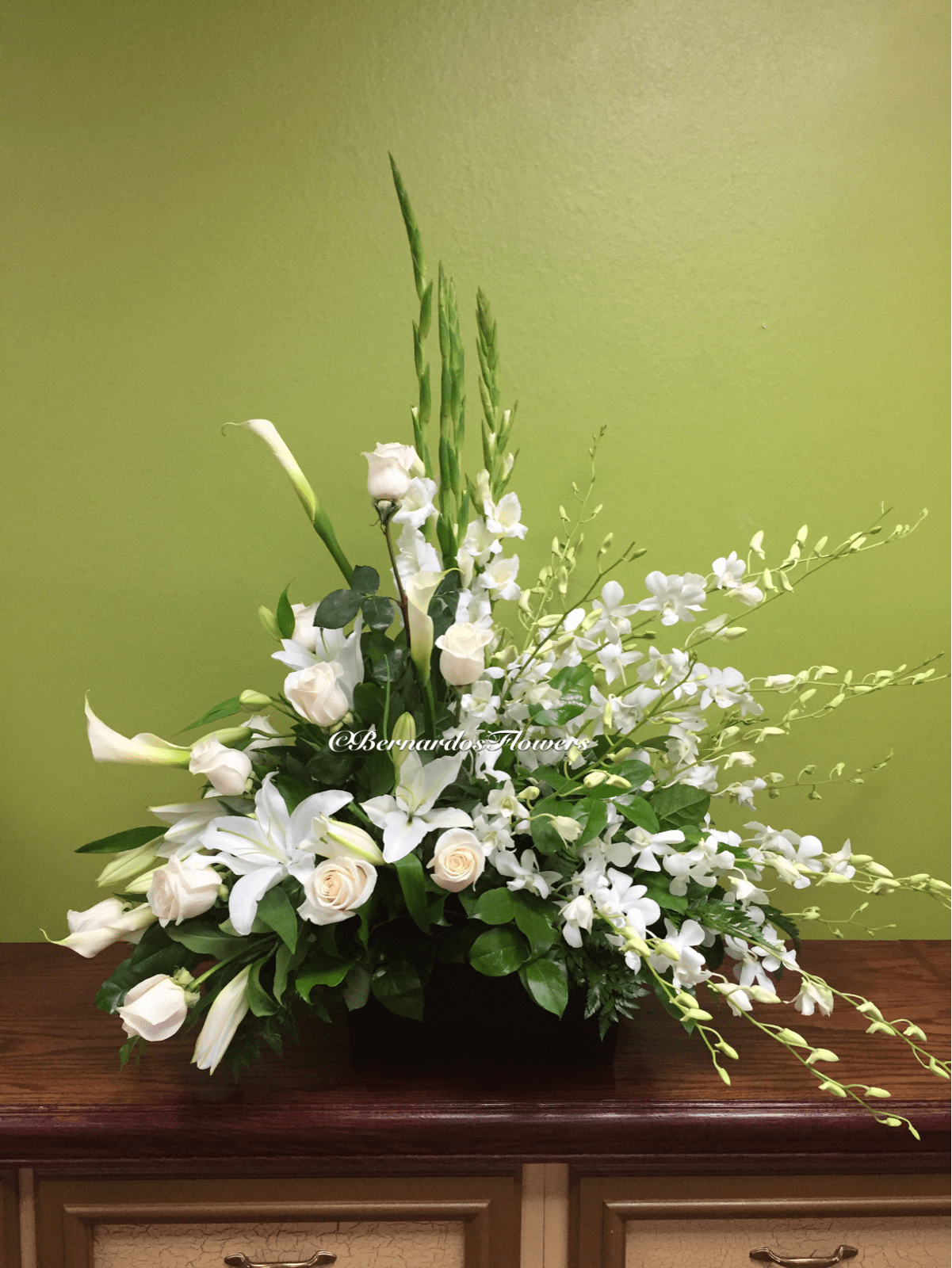 BF-E287 Podium Arrangement - A graceful all-white arrangement featuring roses, lilies, hydrangeas, and orchids. This elegant display offers a heartfelt tribute, symbolizing peace, love, and remembrance.