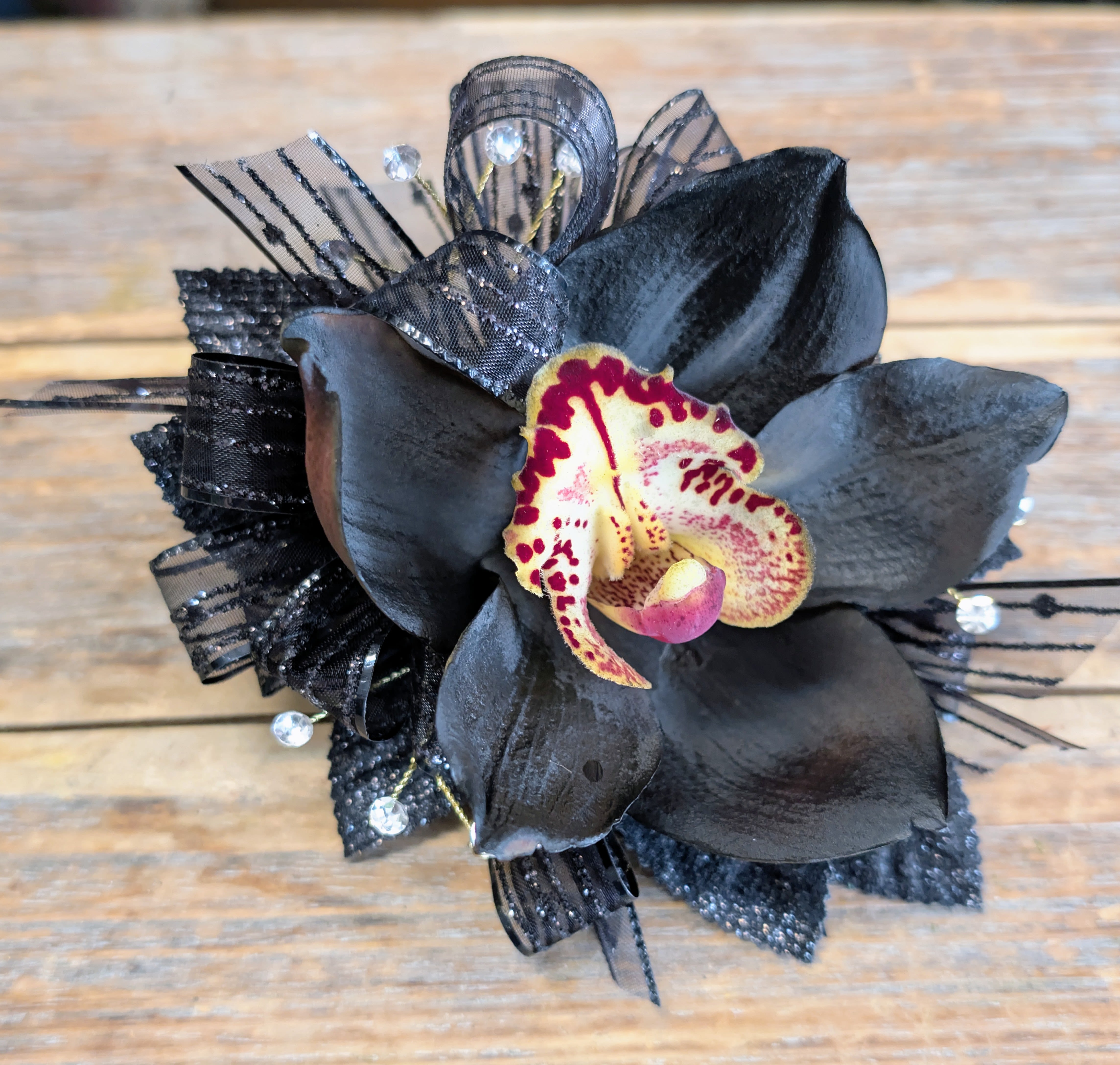 Black Orchid Corsage With Rhinestone Accents - Make an unforgettable statement with our Black Orchid Corsage, a striking blend of sophistication and edge, exclusively from Windmill Floral Expressions in Las Vegas. This design showcases a dramatic black orchid with deep, velvety petals, highlighted by a vivid center of golden yellow and burgundy tones. Accented with shimmering black ribbon, sparkling crystals, and delicate textural details, it’s the perfect accessory for those who want to stand out with elegance and artistry.