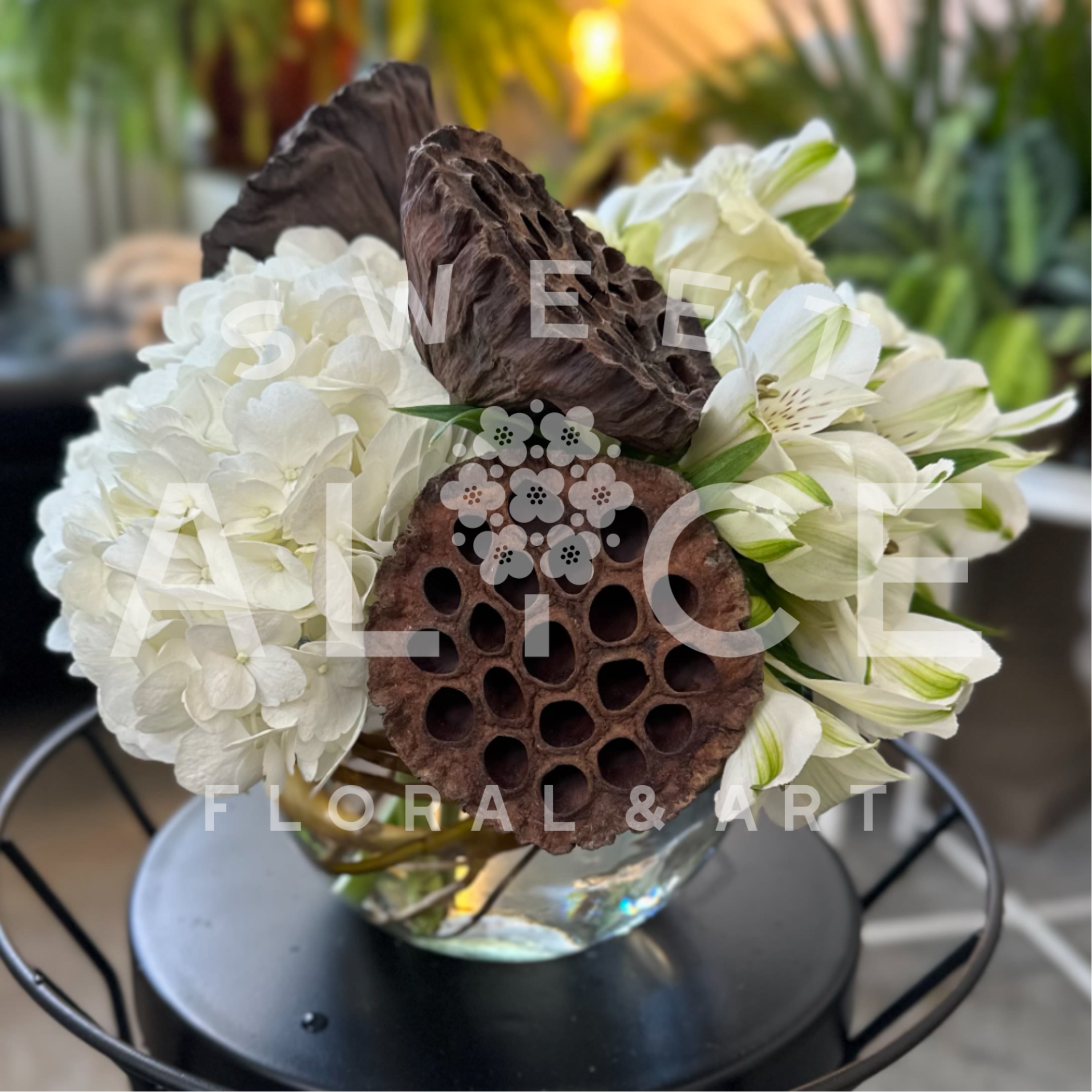 Hoarfrost - Reminiscent of a winters morning, lush white hydrangeas, spray roses and alstroemeria dance with brown lotus pods and curly willow in this elegant centerpiece.   Approximately 9" wide by 9" high.