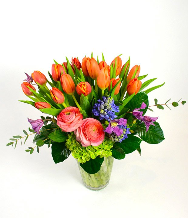 Fresh Squeezed - Orange crush tulips, fragrant hyacinth, peach ranunculus with clematis and hydrangea presented in clear glass. Fresh, vivid and inviting.