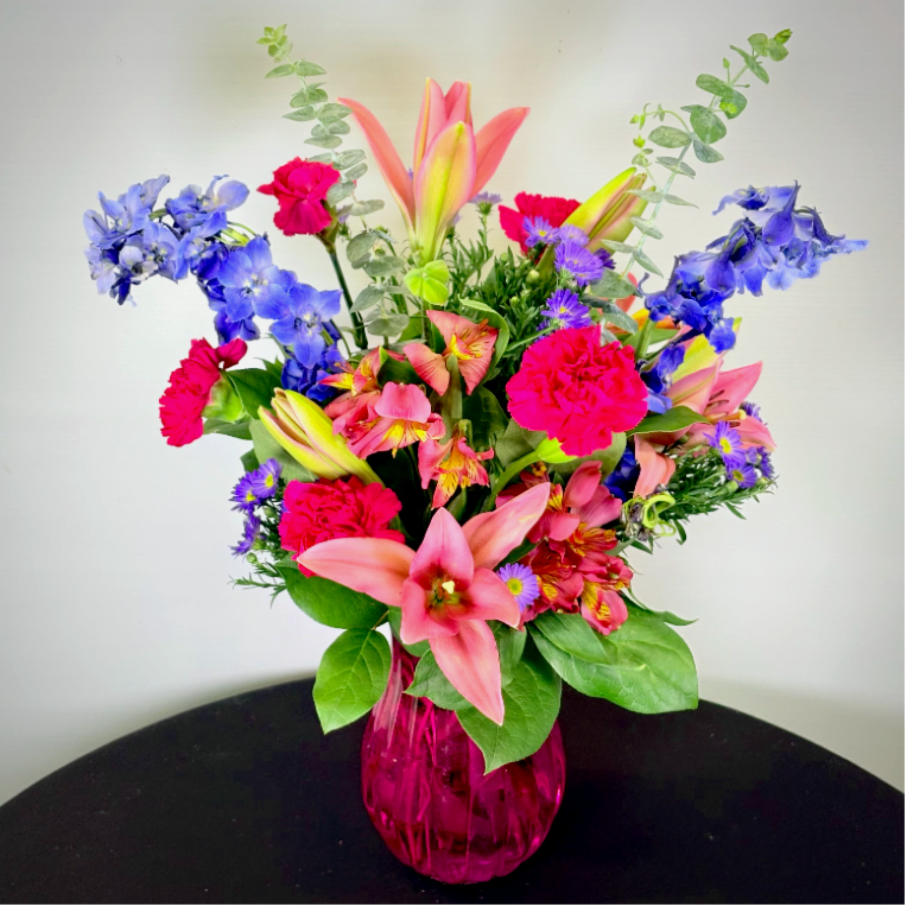 Wild About You - A bold and romantic bouquet featuring pink lilies, lush carnations, and pops of blue delphinium, artfully arranged in a colored glass vase. Full of color and charm, it's a stunning way to say, "I'm wild about you!"
