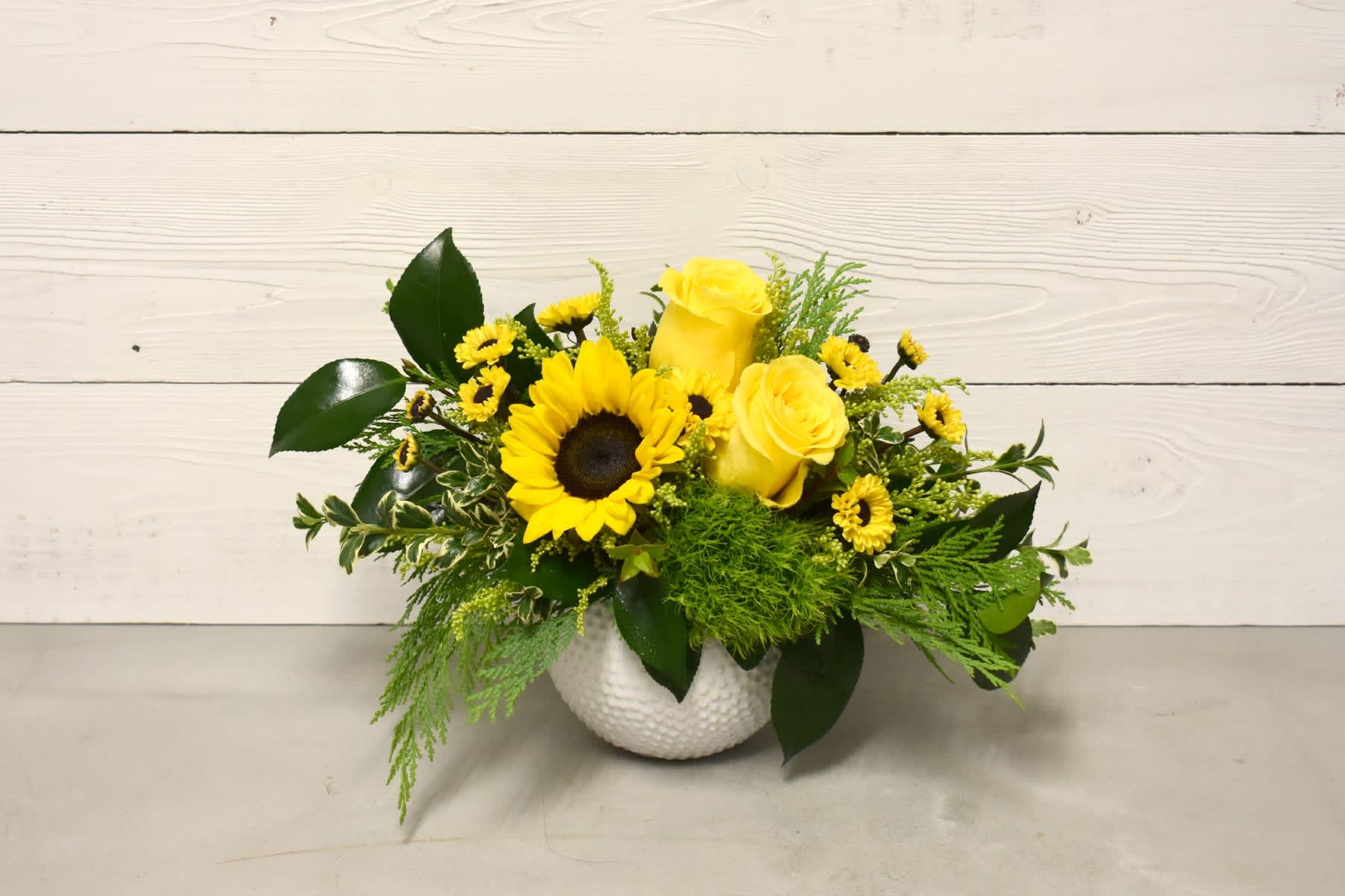 Daphne - This arrangement features a mix of roses, sunflowers, chrysanthemums, dianthus, greens, and filler in a white ceramic vase. 