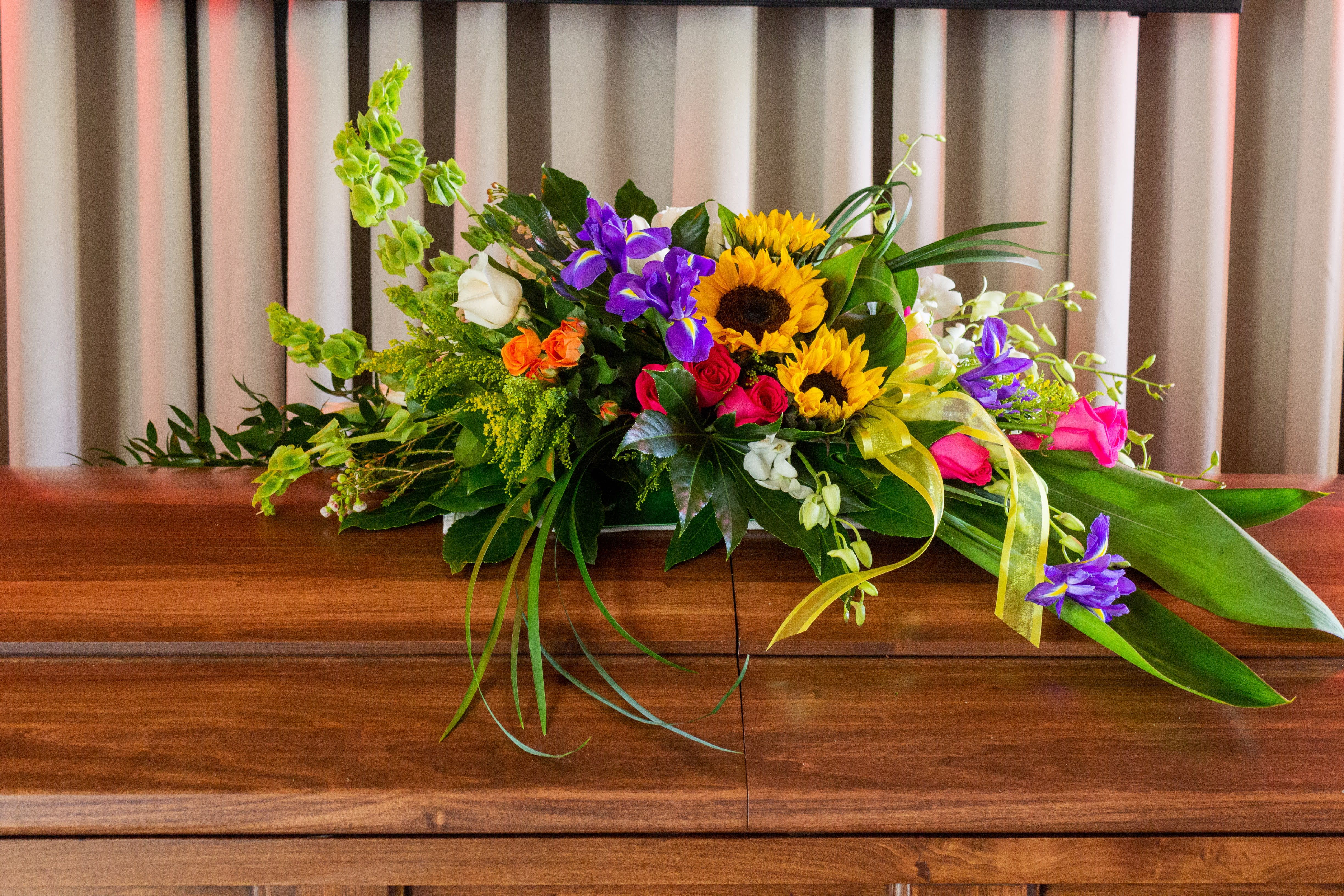 FUNERAL CASKET SPRAY - Morning Rays 103 - This is a wonderful option for a funeral service or church. This casket can be designed with a saddle that rests over approximately half of the casket for an open casket or the full casket for a closed casket service. This lovely mix of yellows, pinks, purples, and oranges, with sunflowers, roses, dendrobium orchids, and iris and is a great way to add a splash of color to your service.   **This is a piece for a funeral or memorial service. Please place the order at least 2 days prior to the service to guarantee a certain arrangement. Anything sooner and we cannot guarantee it will match the picture. These pieces can be customized to the color of your choice. The flowers may vary depending on availability of the flowers, special requests and quality of the flowers we have in stock. If you have a special request please add it to our special instructions when placing your order. If you have any questions please don’t hesitate to call. 714 289 1123**